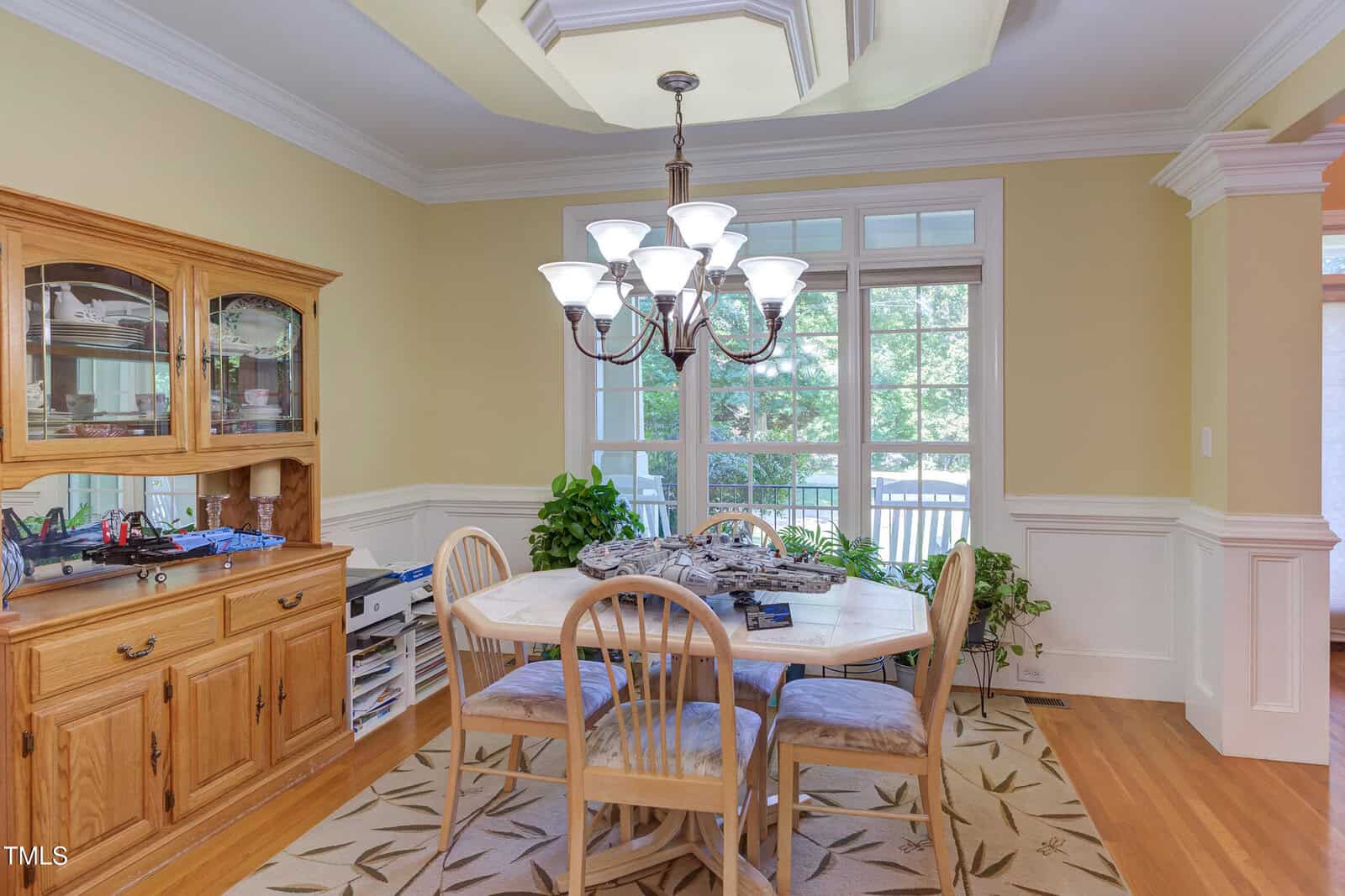 A dining room with a wooden table and four chairs, a chandelier above, a wooden hutch on the left, houseplants by the windows, and large windows letting in natural light.