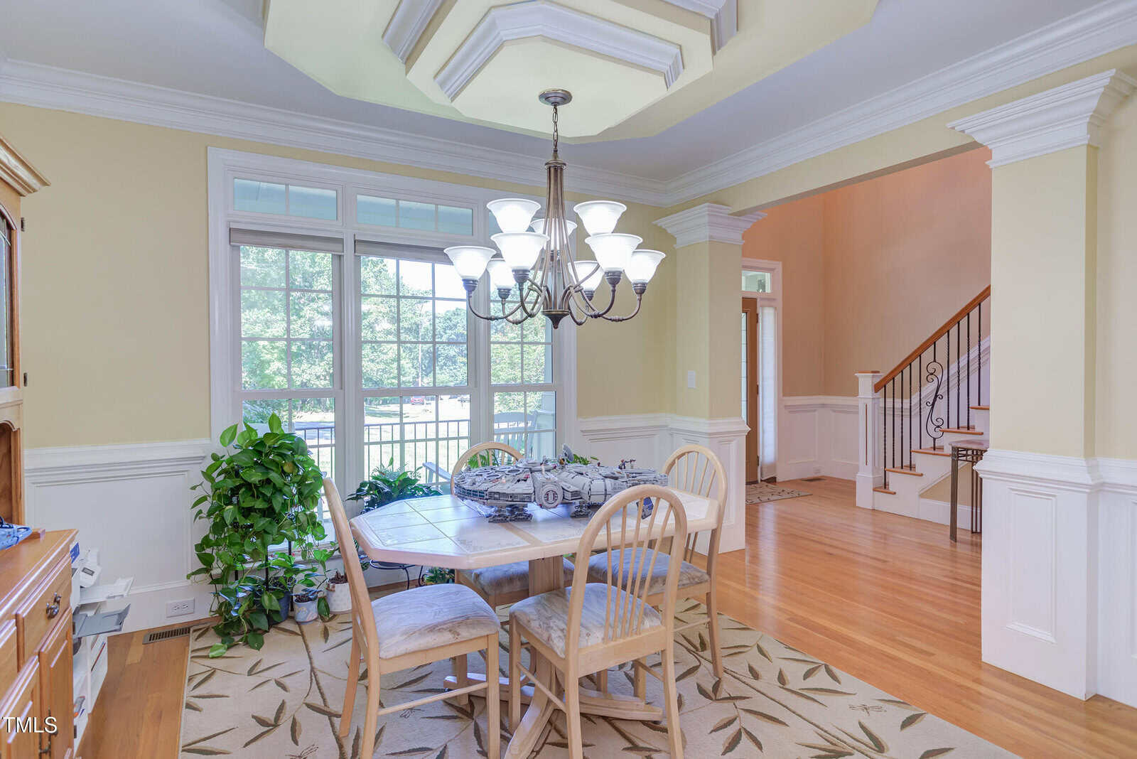 A bright dining room with a wooden table, four chairs, a chandelier, large windows, a green plant, and a staircase visible through an open doorway.