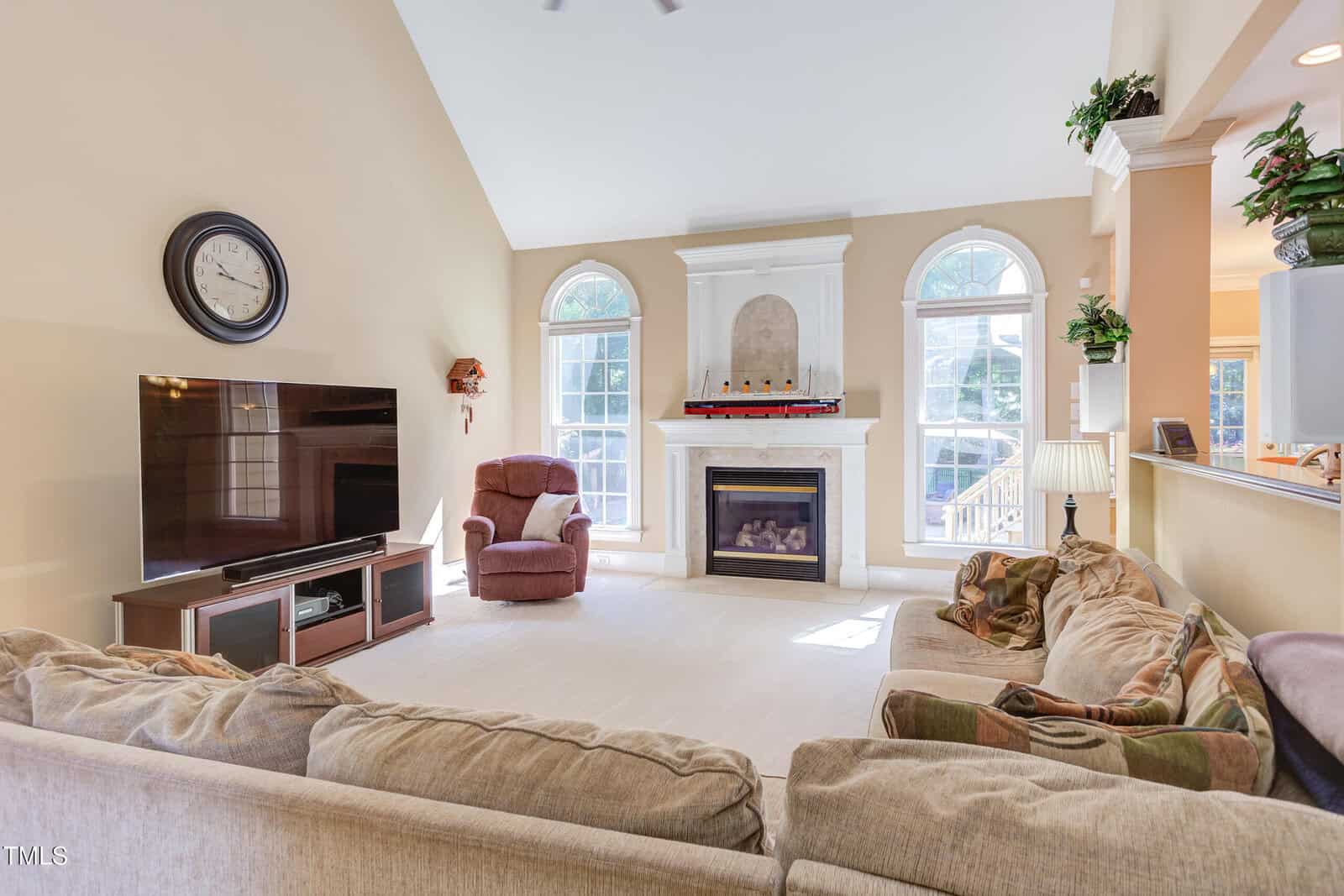 Bright living room with a large sectional sofa, TV, wall clock, fireplace, and three tall windows. Armchair near the windows and several potted plants decorate the space. Light walls and carpet create an airy feel.