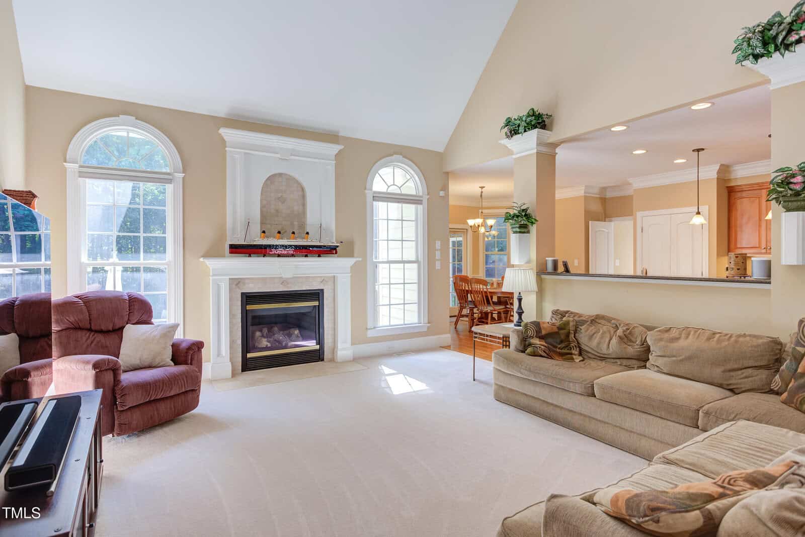 Bright living room with tan walls, large windows, a fireplace, a beige sectional sofa, a red recliner, and an open view into a dining area and kitchen with wood cabinets and pendant lights. Greenery is placed above cabinets.