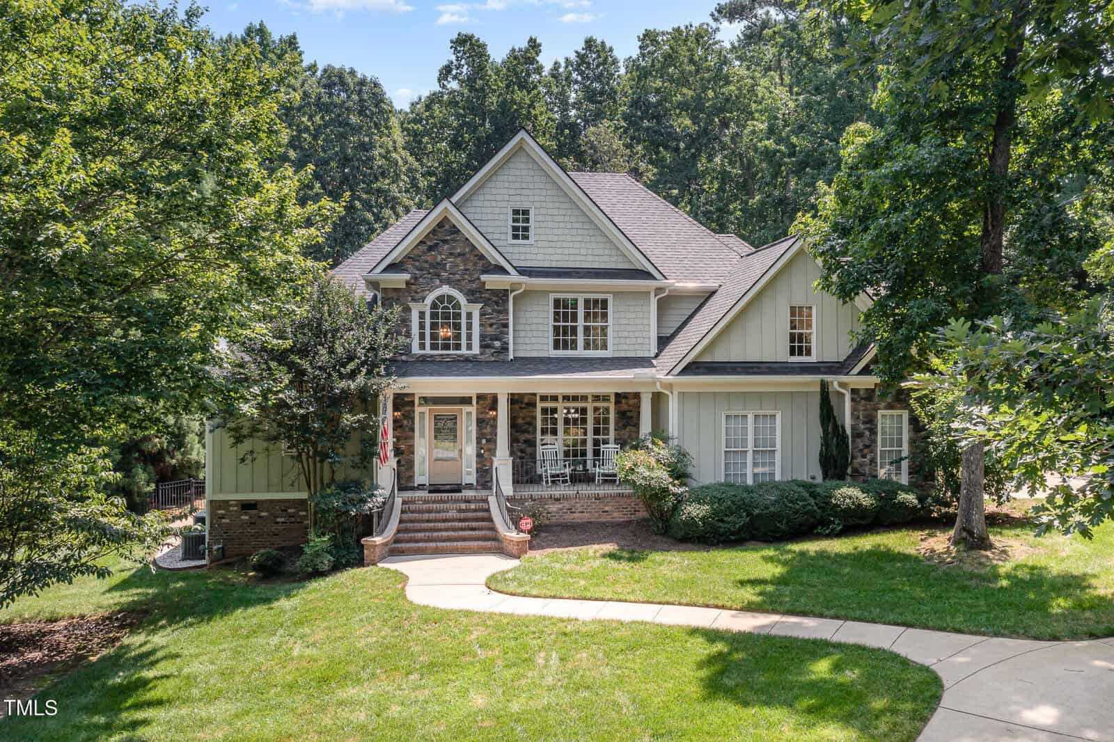 A large two-story house with light-colored siding, stone accents, a front porch with white railings, and multiple gables, surrounded by trees and greenery, with a curved walkway leading to the entrance.
