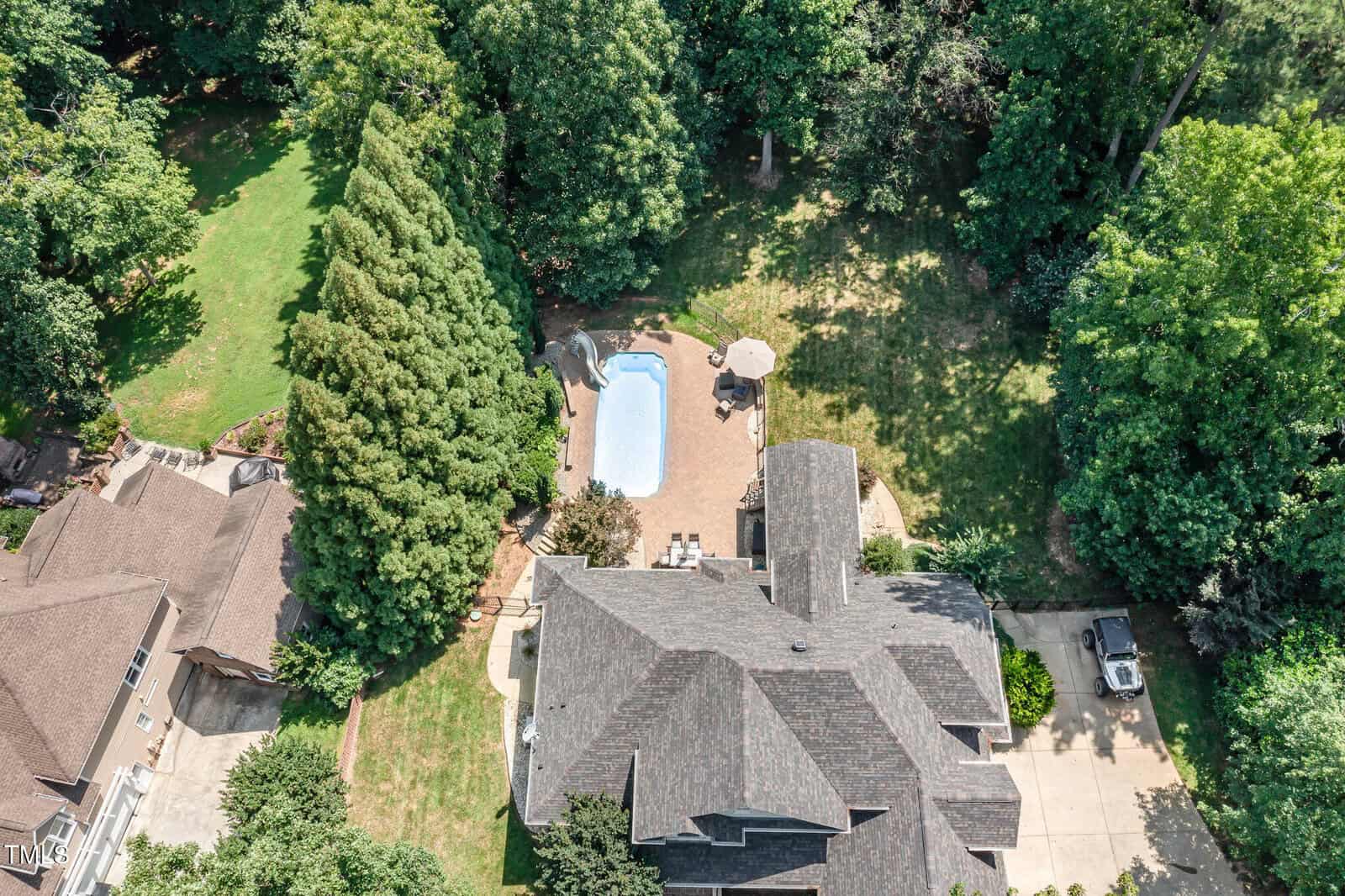 Aerial view of a house with a shingle roof, a backyard pool surrounded by a brick patio, lounge chairs, a table with an umbrella, tall trees, and a driveway with parked vehicles.
