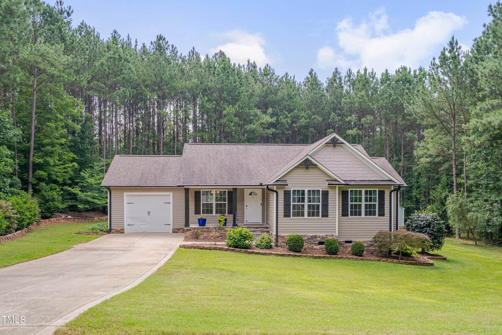 A single-story house at 1210 Bayview Court, Franklinton, with light-colored siding, an attached garage, a covered front porch, and large windows sits on a grassy lawn surrounded by landscaping and tall pine trees.