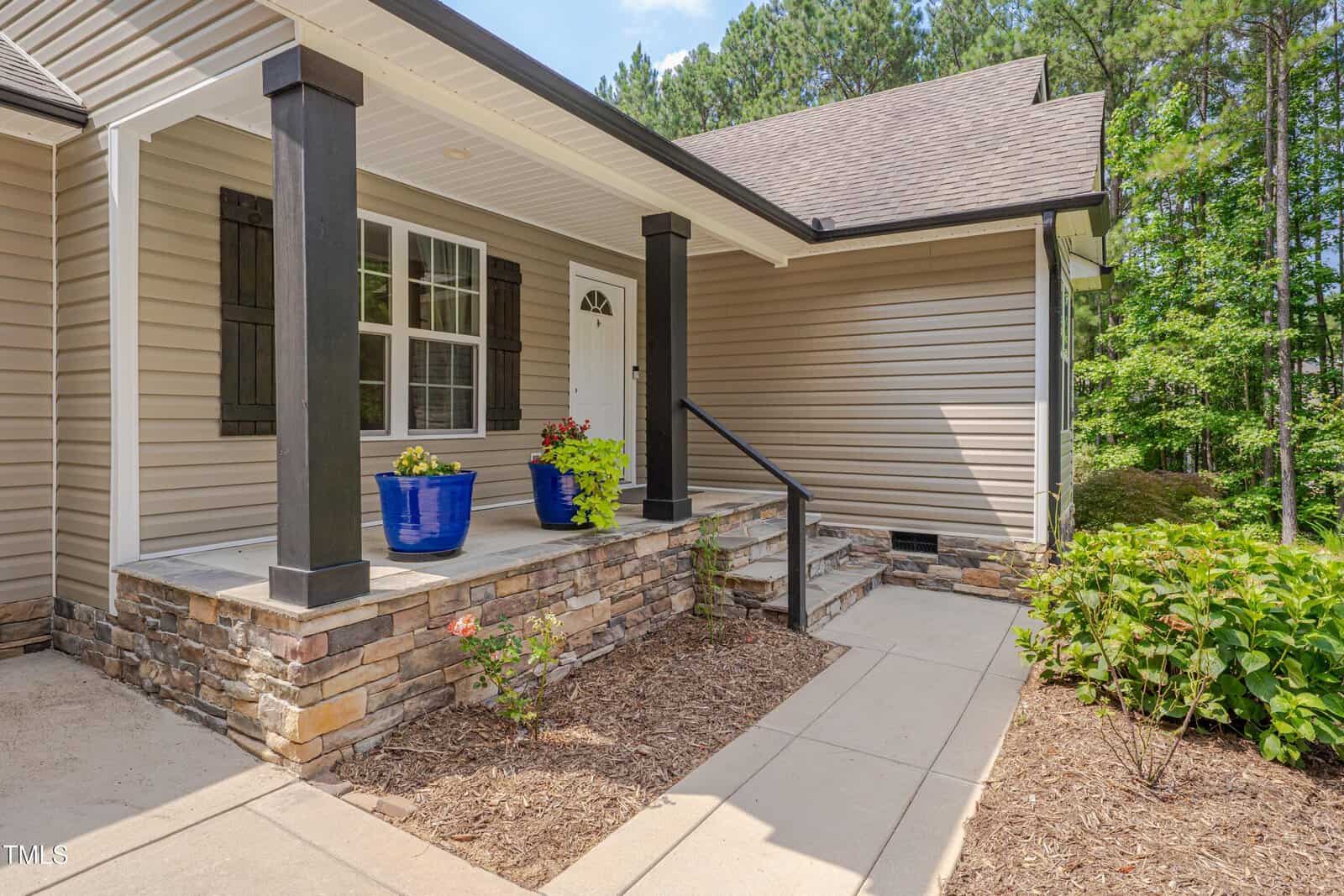 A tan house with stone accents features a covered front porch supported by black columns. Blue planters with green plants sit near a white front door. A ramp and sidewalk lead to the entrance. Shrubs are planted nearby.