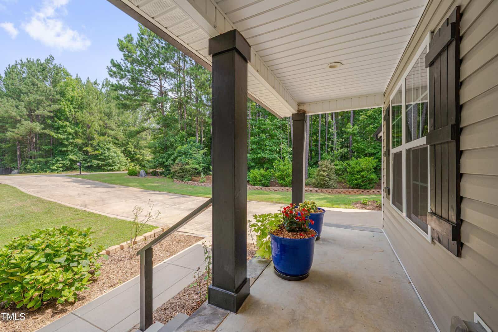A view from a covered front porch with black columns, beige siding, and two blue planters with flowers. The porch overlooks a driveway, green lawn, and trees in the background.