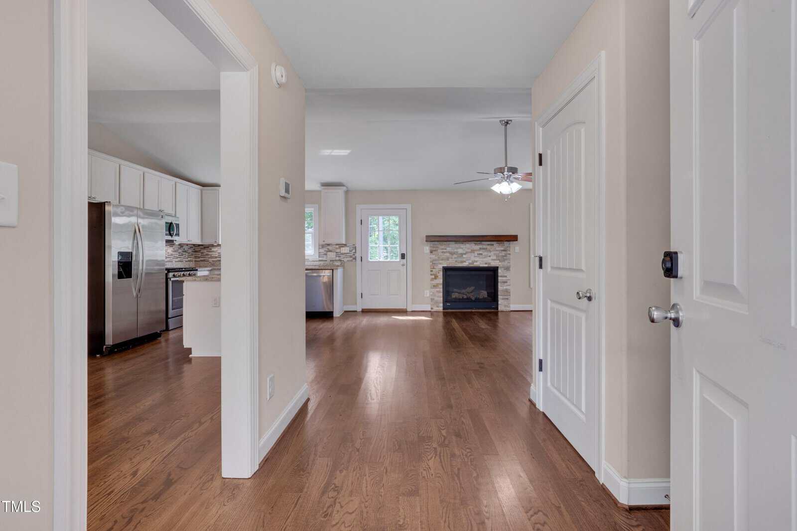 View from the entryway of a house showing a hallway with wood flooring, leading to a kitchen with white cabinets and stainless steel appliances on the left, and a living room with a fireplace straight ahead.