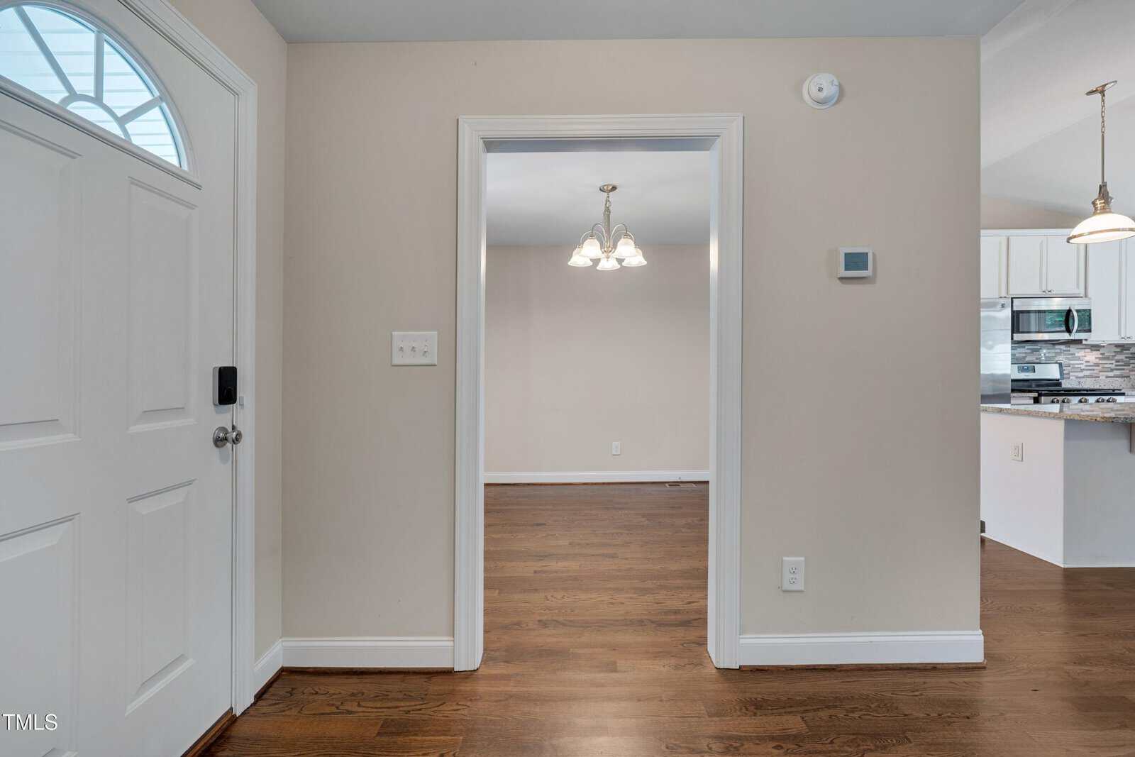 View from an entryway looking into a room with hardwood floors, beige walls, a hanging light fixture, and a doorway leading to a kitchen with stainless steel appliances and white cabinets.