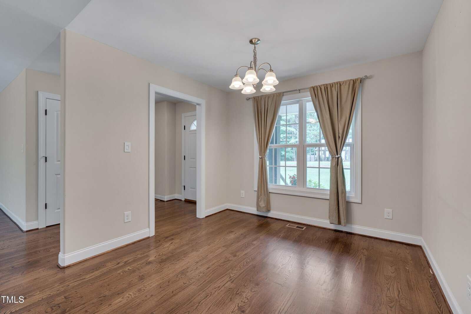 A small empty room with light beige walls, hardwood floors, a window with brown curtains, and a ceiling light fixture. There is an open doorway leading to an adjacent space.