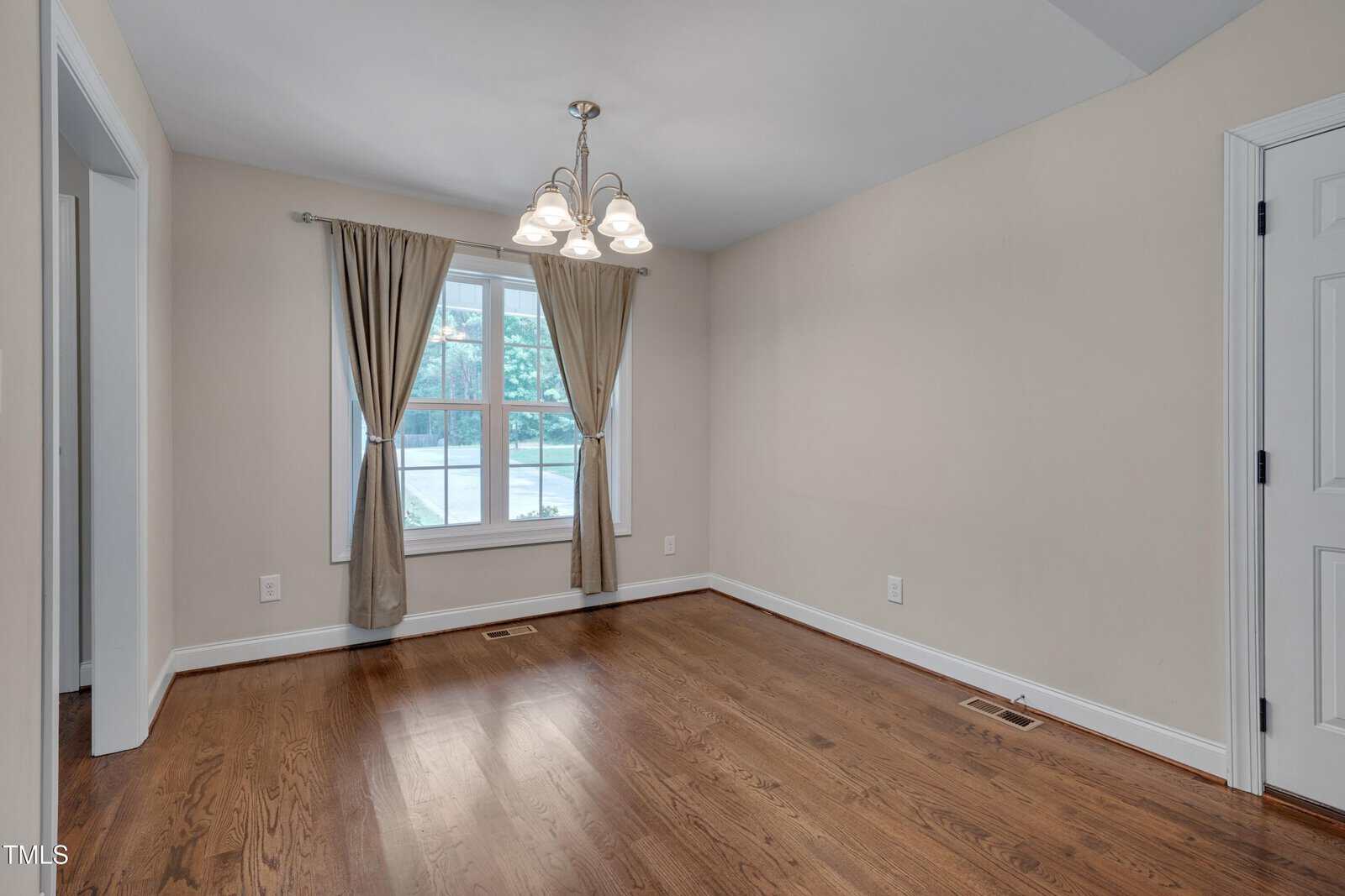 A small, empty room with hardwood floors, beige walls, a window with brown curtains, a ceiling light fixture, and white baseboards. There is a white door on the right side of the room.