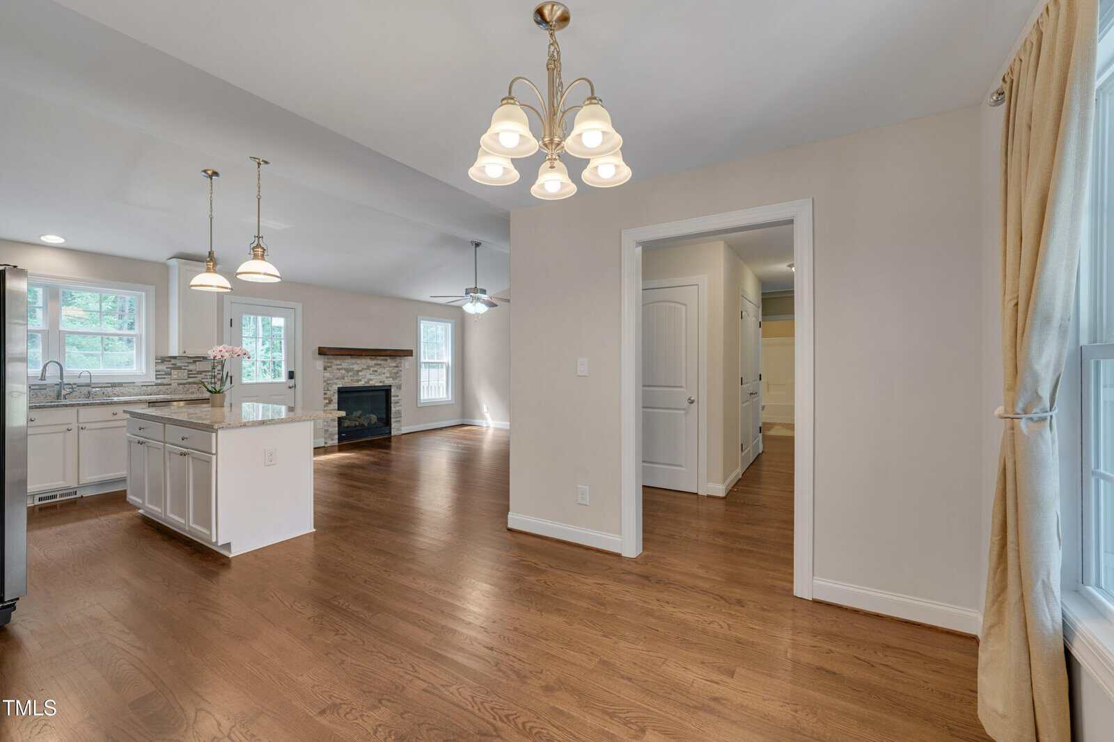 Open-concept kitchen and dining area with hardwood floors, white cabinets, pendant lighting, a fireplace in the living space, and large windows with beige curtains allowing natural light in.