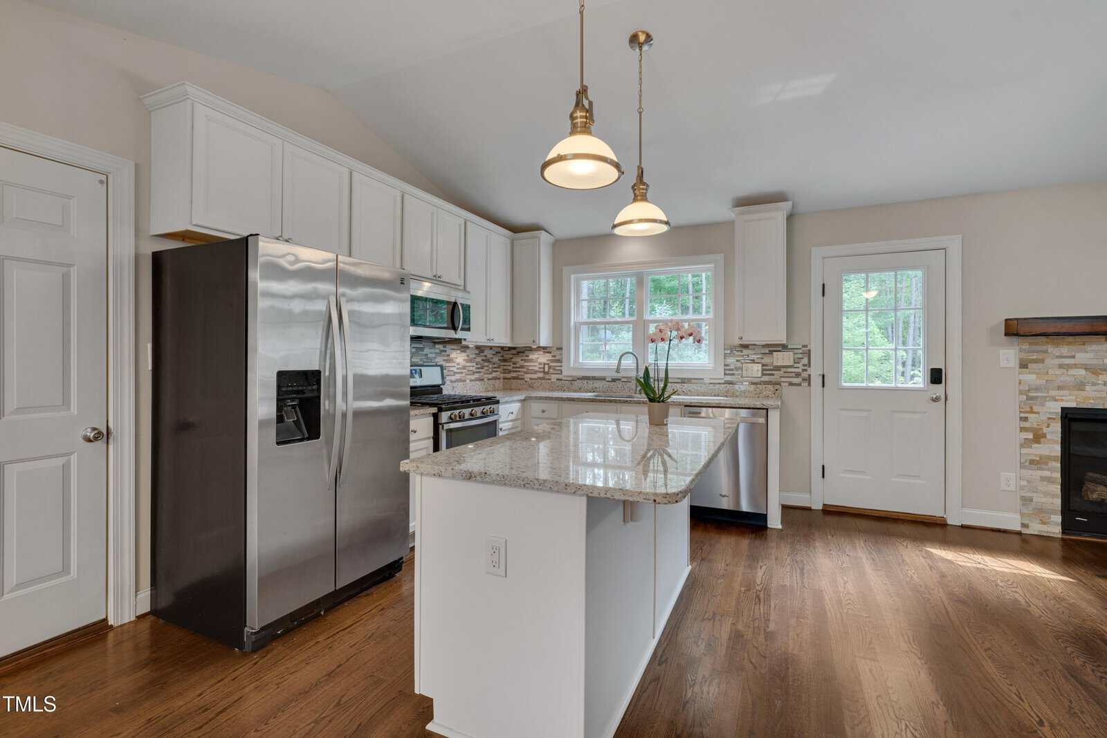 Modern kitchen with stainless steel appliances, white cabinets, granite countertops, a center island, wooden floors, pendant lights, and a windowed door leading outside.