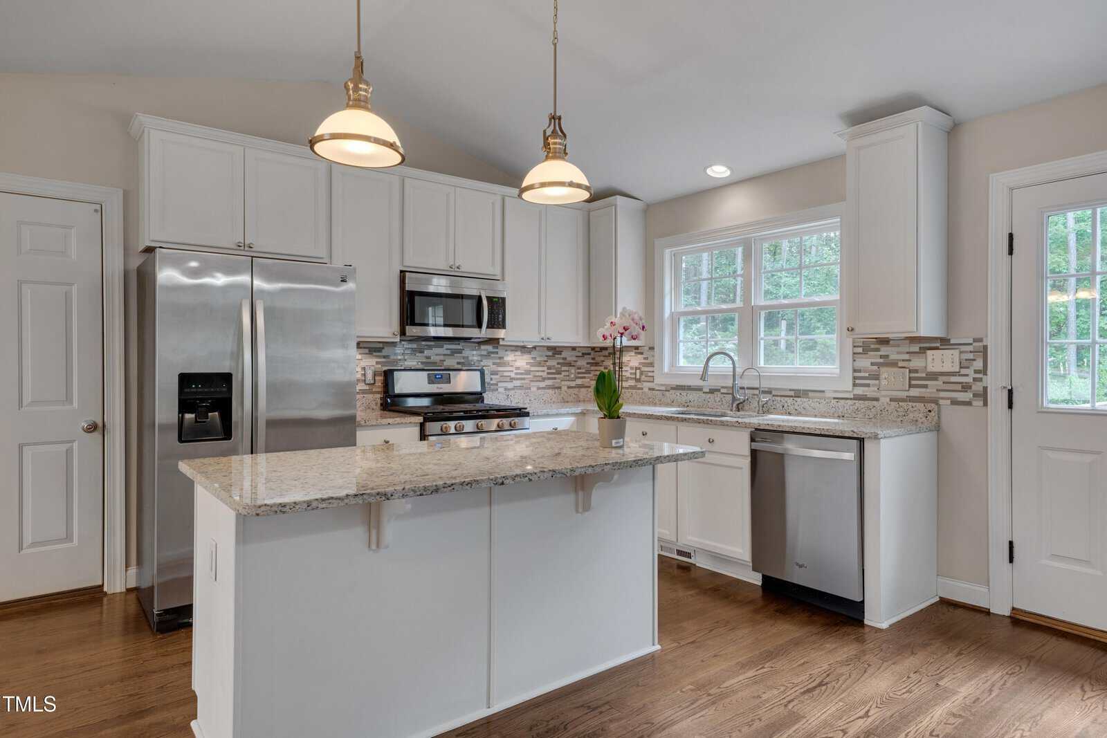 Modern kitchen with white cabinets, stainless steel appliances, granite countertops, a center island with two pendant lights above, tile backsplash, wood flooring, and a window over the sink with natural light.