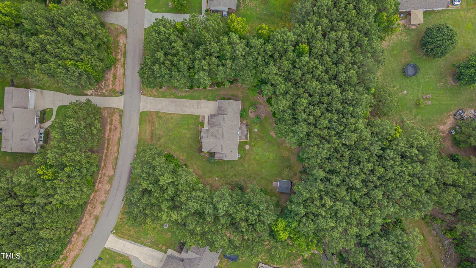 Aerial view of a suburban neighborhood with houses, driveways, and surrounding trees. A curved road runs through the image, with dense wooded areas between and behind the homes. A trampoline is visible in a backyard.