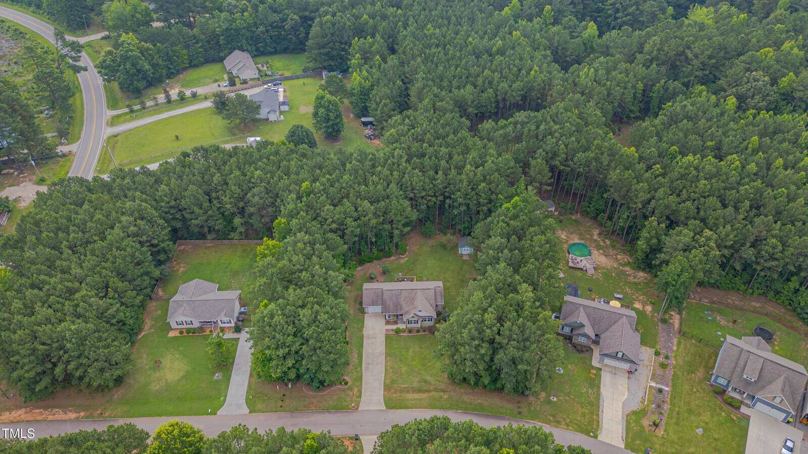 Aerial view of a suburban neighborhood with three houses along a street, large front yards, driveways, and dense trees surrounding the properties; additional houses and roads are visible in the background.