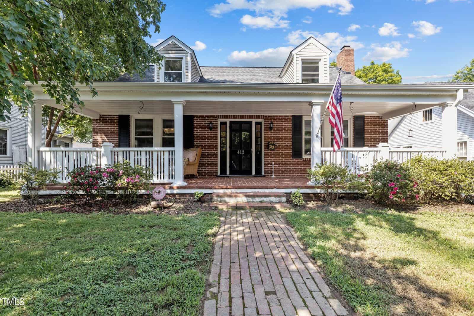 A brick house at 412 Coggeshall Street in Oxford features a large covered front porch with white railings and an American flag. A brick walkway winds through landscaped bushes and flowers, leading to the inviting front steps under a blue, cloud-streaked sky.