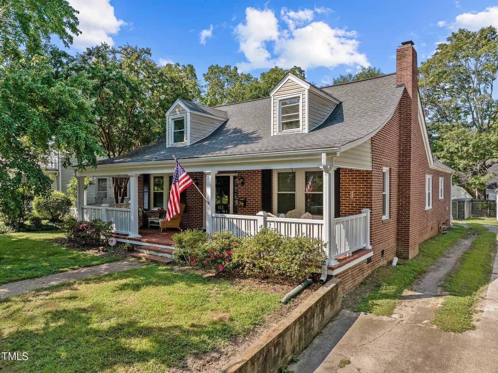 A brick house with a covered front porch, two dormer windows, and an American flag. The porch has white railings and outdoor furniture, with bushes and a green lawn in front. Trees and a blue sky are in the background.