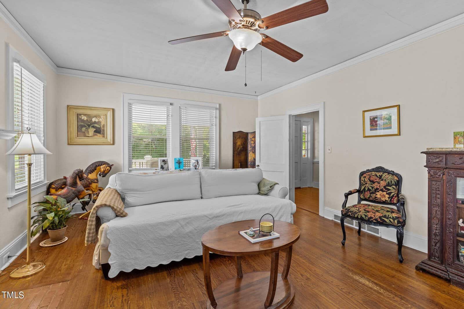A living room with a white sofa covered by a slipcover, a patterned armchair, a wooden coffee table with books, a ceiling fan, a fireplace mantel, potted plants, framed art, and hardwood floors.