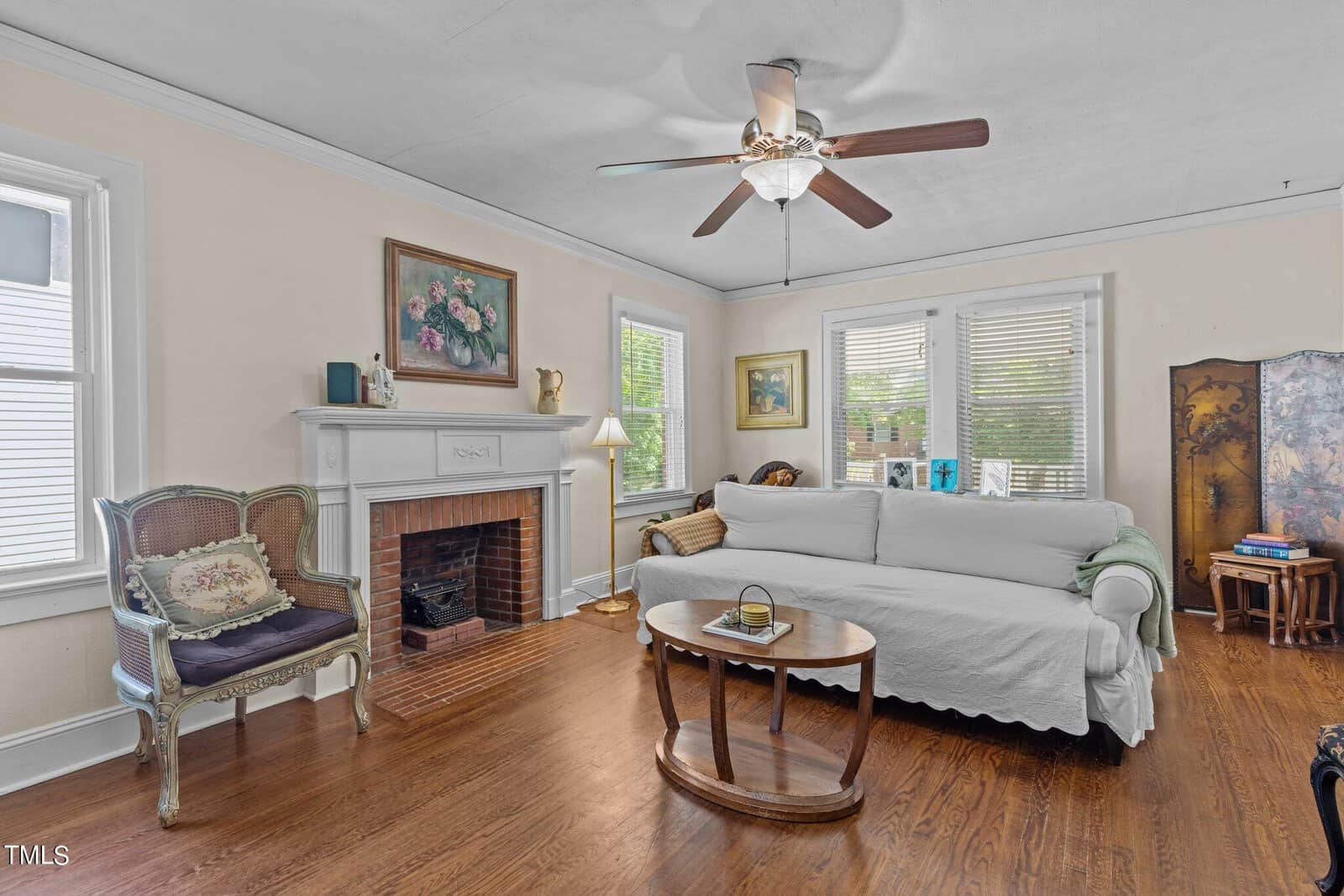 A living room features a white sofa with a white cover, a wooden coffee table, a decorative armchair, a fireplace with artwork above it, wood floors, and several windows letting in natural light.
