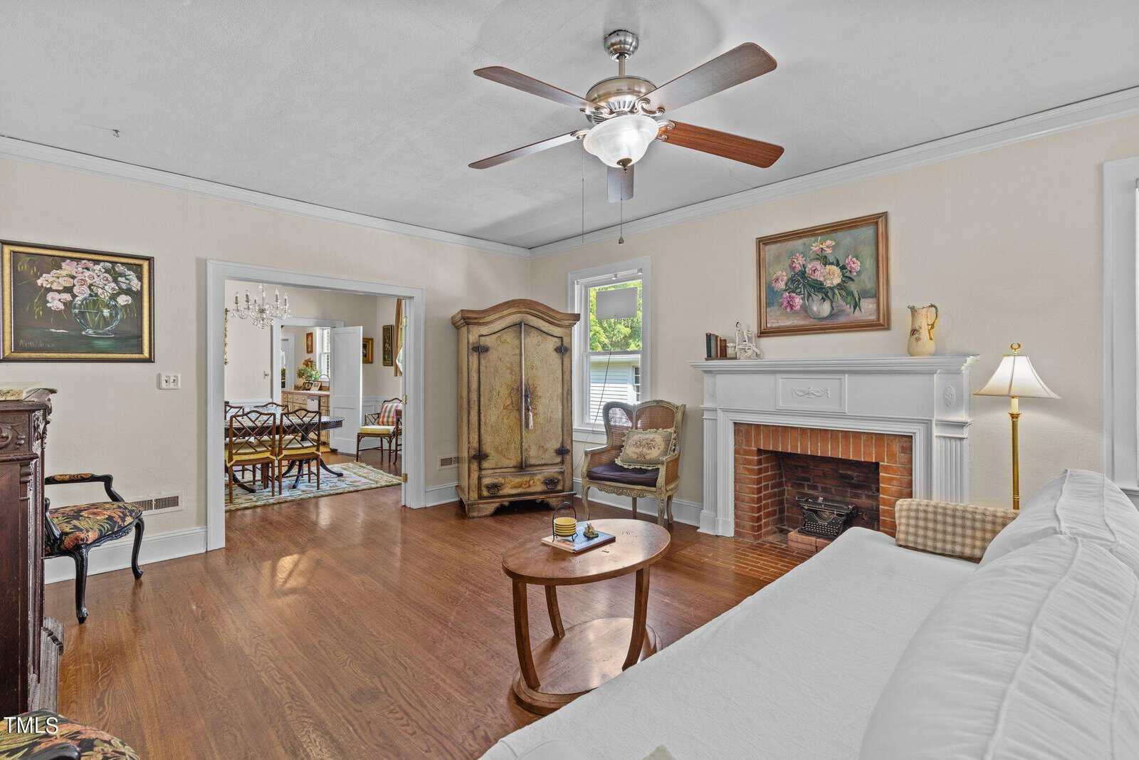 A cozy living room with wood floors, a white couch, a round coffee table, a fireplace, an armoire, and ceiling fan. Art and flowers decorate the walls. A dining area is visible through an open doorway.