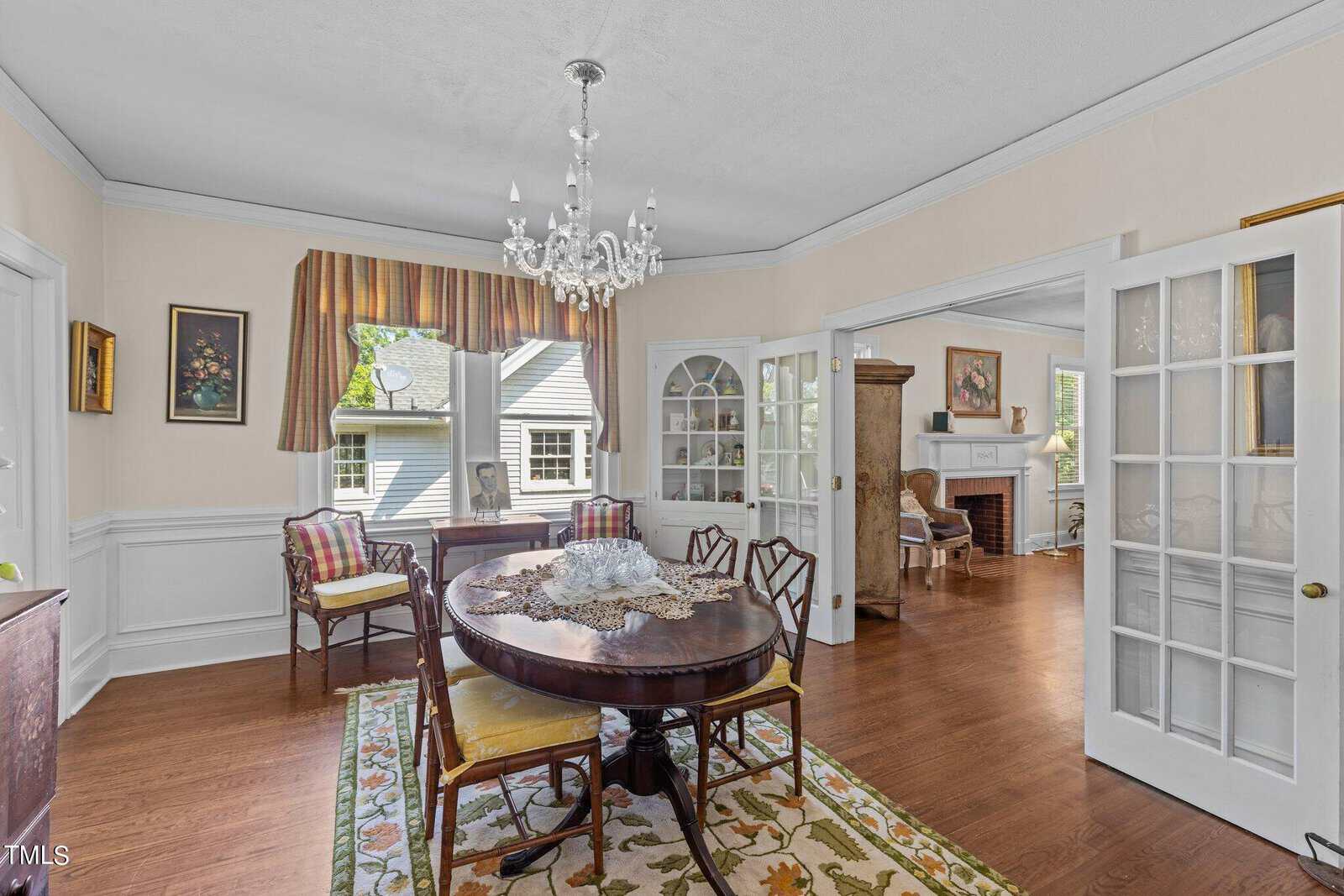 Bright dining room with round table, chandelier, floral rug, and open French doors leading to a living room with fireplace and large windows. Wall art and a window with striped valance decorate the space.