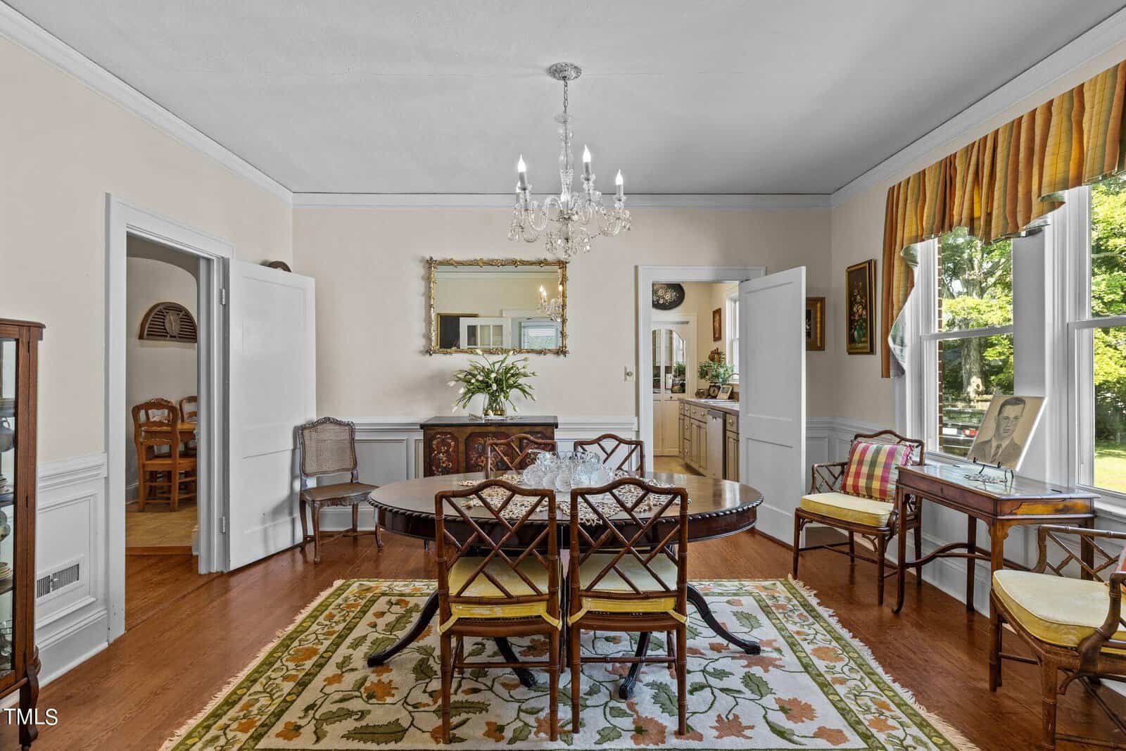 Bright dining room with a round wooden table, six chairs, a floral rug, chandelier, side tables, and large windows with striped curtains overlooking a green yard.