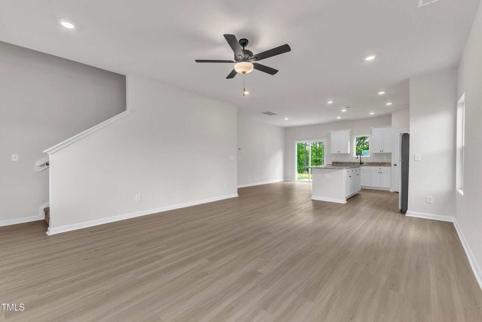 A spacious, empty open-concept living area with light wood flooring, white walls, a ceiling fan, a staircase to the left, and a modern kitchen with an island and white cabinets in the background.