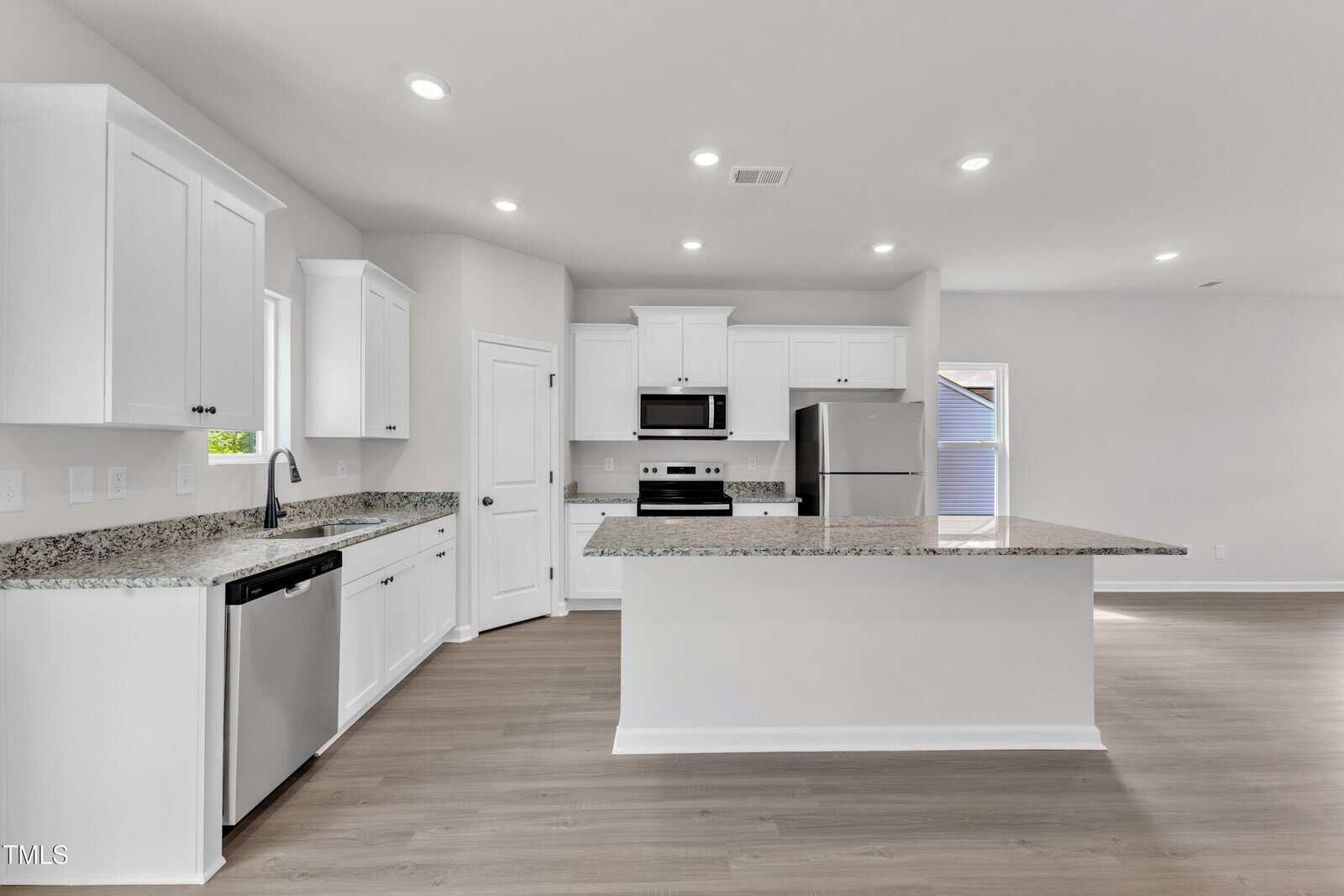 Modern kitchen with white cabinets, stainless steel appliances, granite countertops, island with sink, light wood flooring, and recessed lighting. A pantry door and two windows provide natural light.
