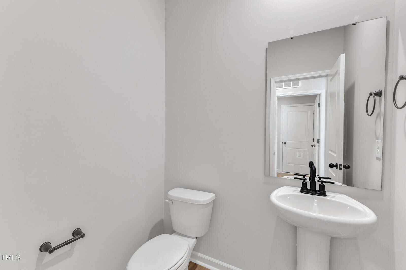 A small bathroom with a white toilet, a white pedestal sink, a rectangular mirror above the sink, a wall-mounted towel ring, and a toilet paper holder. The walls are light gray.