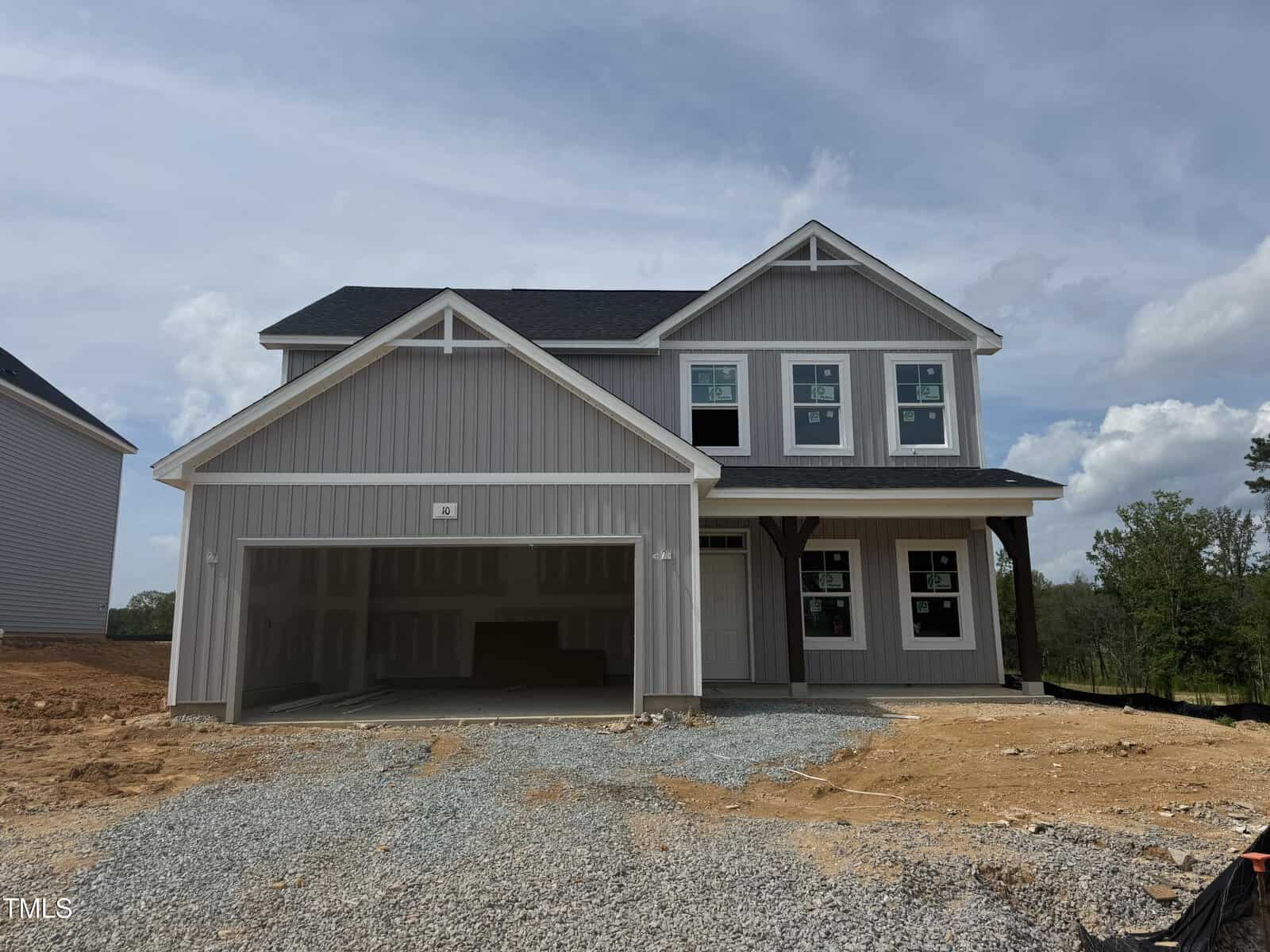 A newly constructed two-story gray house with white trim, an attached two-car garage, and several windows. The garage door is open, and the driveway is unpaved with gravel and dirt in front. Trees are visible in the background.