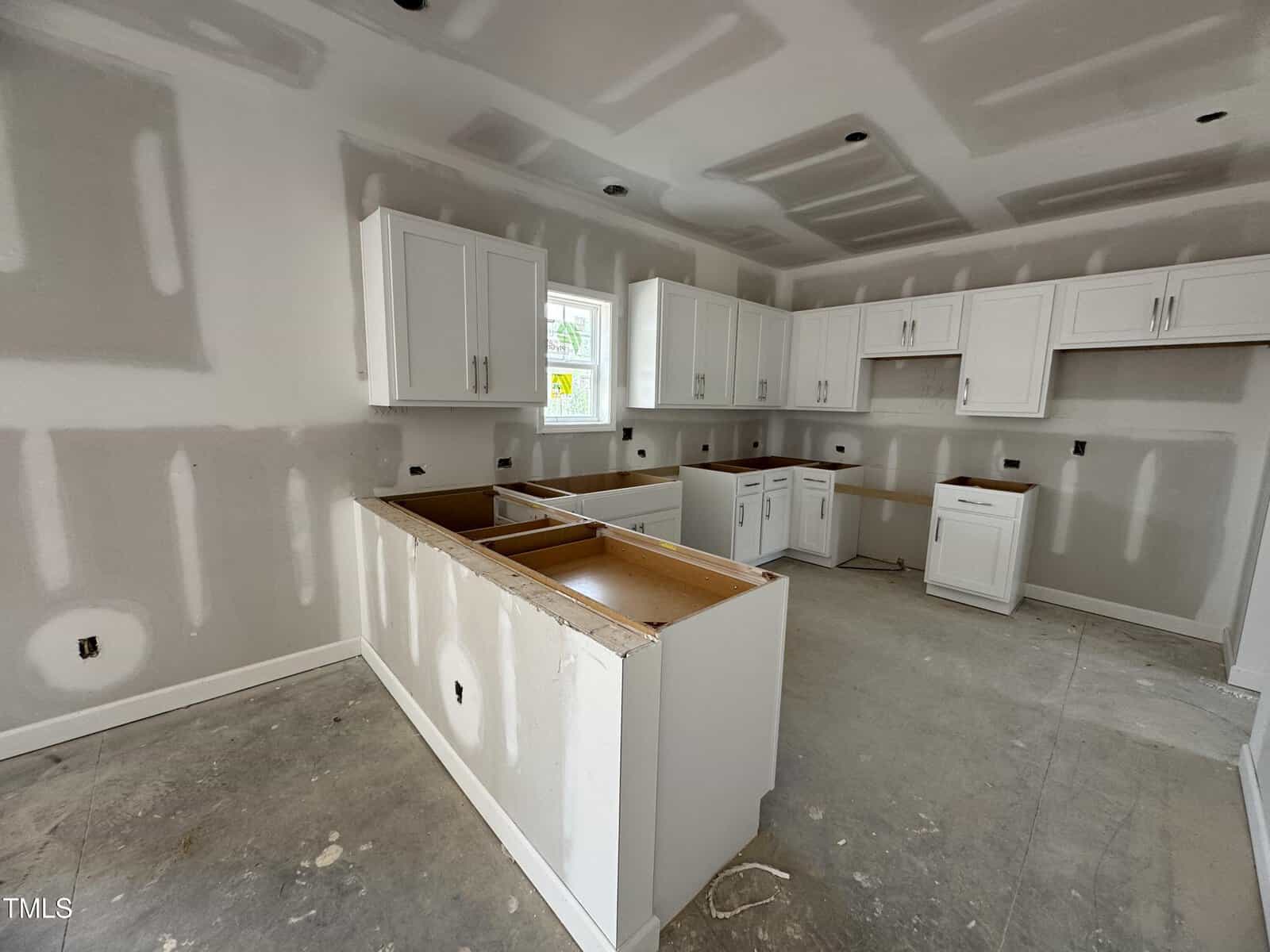 Partially finished kitchen with unpainted drywall, white cabinets installed, and unfinished countertops. The floor is bare concrete, and there are wires and outlets visible in the walls. Natural light enters through a window.