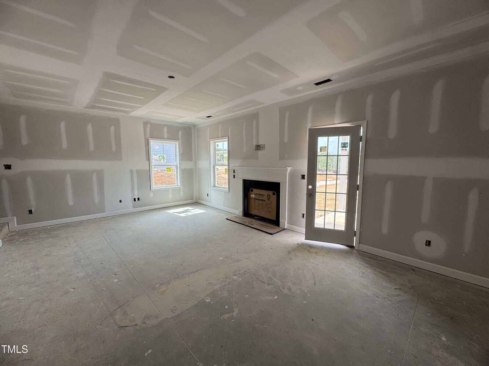 A room under construction with unfinished drywall, two windows, a fireplace with a cardboard cover, a door with glass panes, and a bare floor. Sunlight is coming through the windows and door.