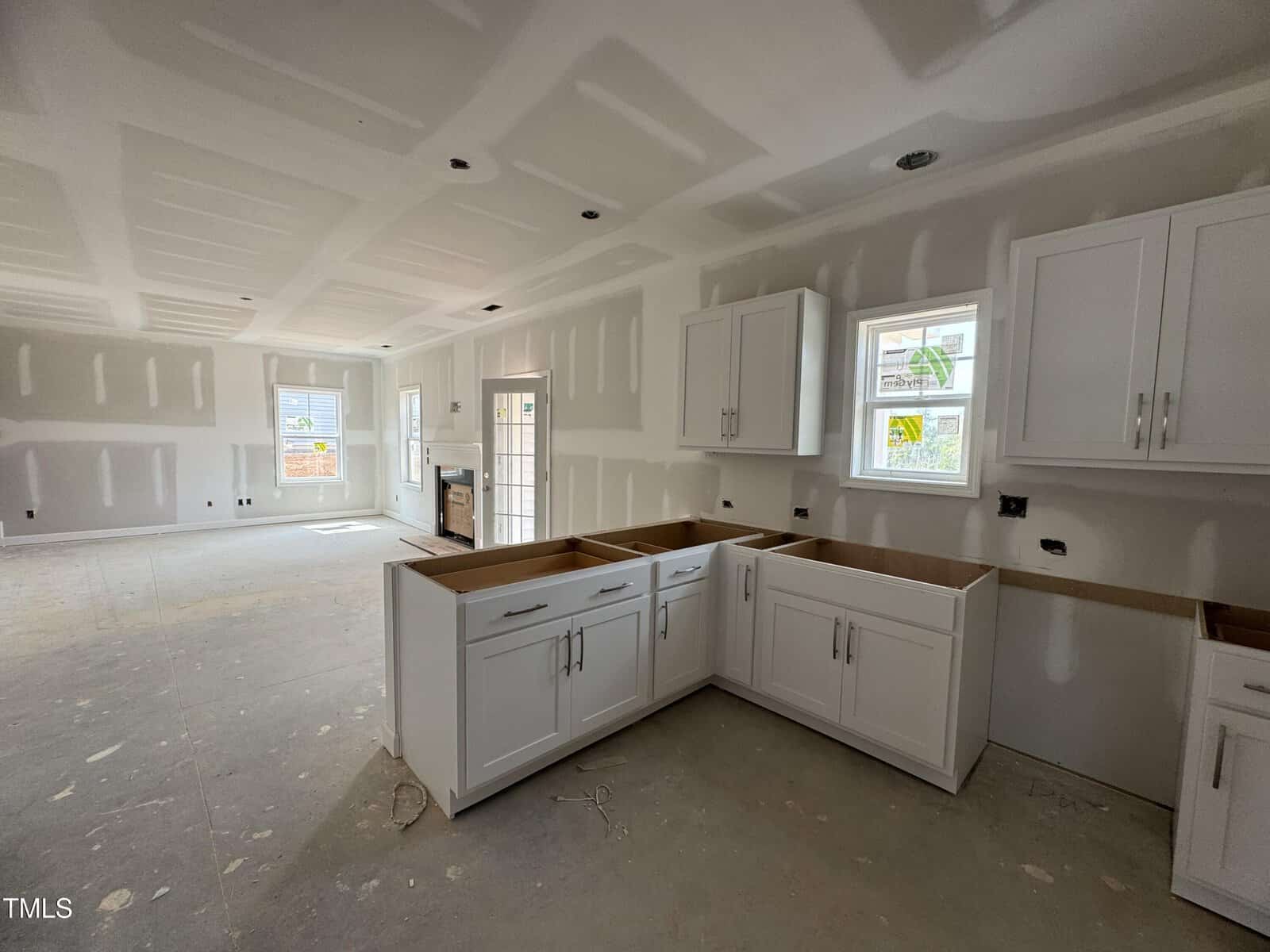 Unfinished kitchen and living area with white cabinets, unpainted drywall, and no flooring. Windows let in natural light; there are no appliances or fixtures installed yet. Construction appears to be in progress.