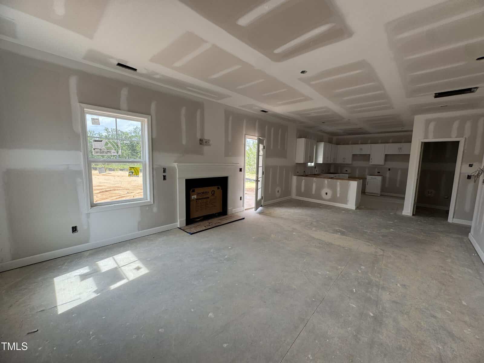 Unfinished living area and kitchen in a new house. Drywall is up with visible seams and patches. Fireplace is installed but not finished. No flooring or paint. Sunlight enters through windows and a glass door.