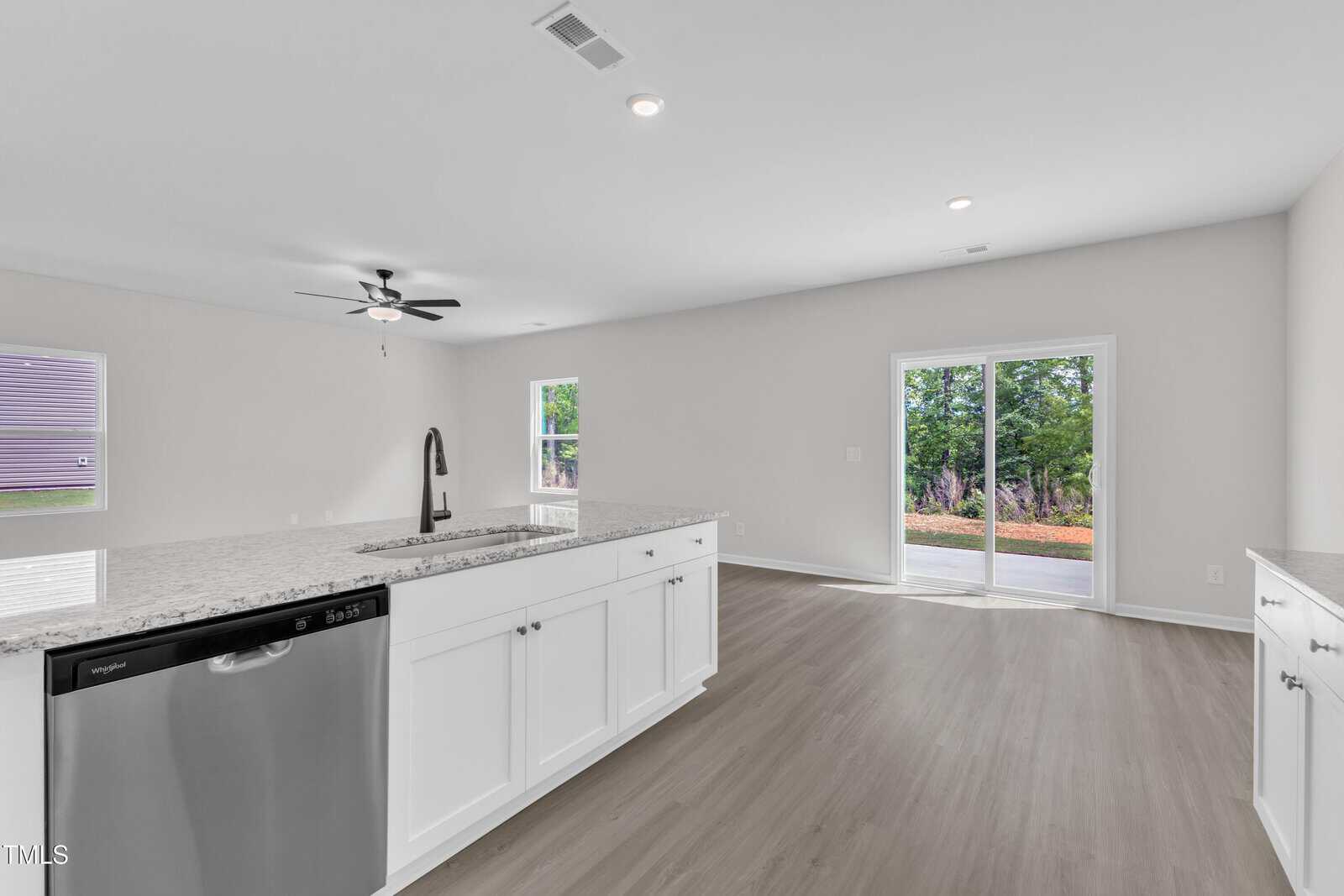 A modern kitchen with white cabinets, granite countertops, stainless steel appliances, an island with a sink, light wood flooring, a ceiling fan, and sliding glass doors leading outside.