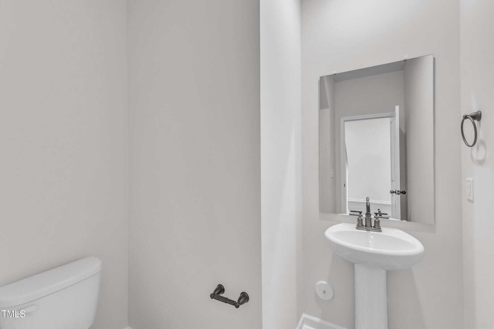 A minimalist bathroom with a white toilet, pedestal sink, mirror, and towel ring on a light-colored wall. There is a black toilet paper holder and a door reflected in the mirror.