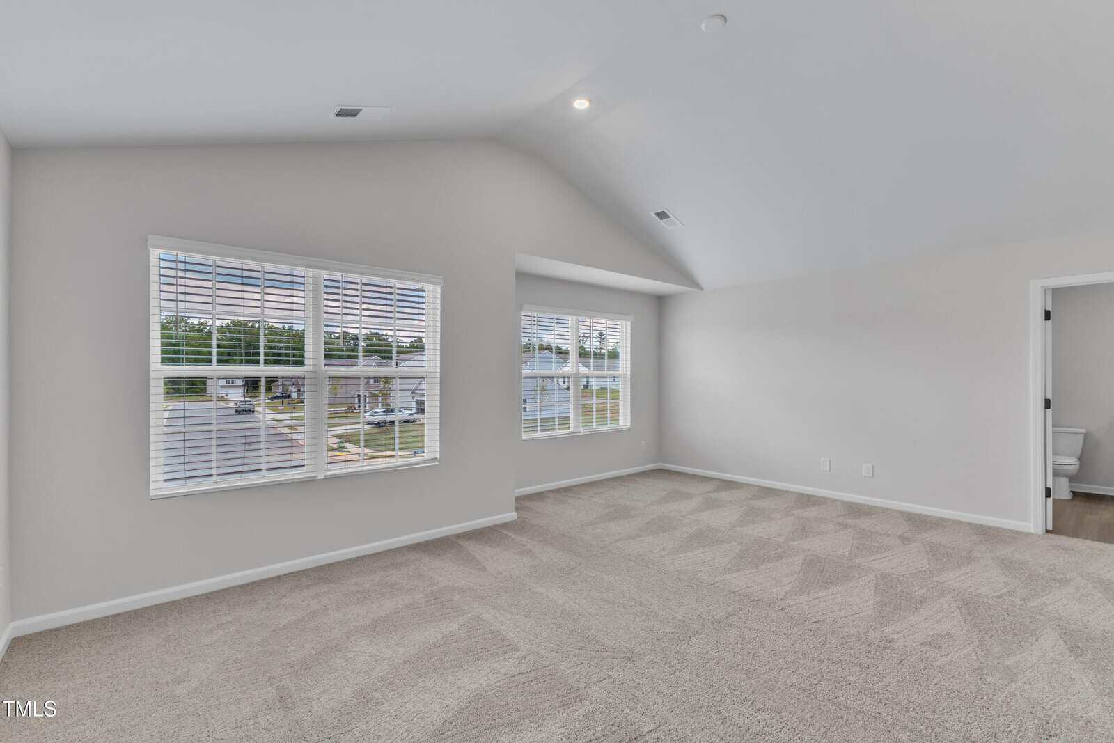 An empty room with light carpet, light gray walls, a vaulted ceiling, two large windows with blinds, and an open doorway leading to a bathroom.