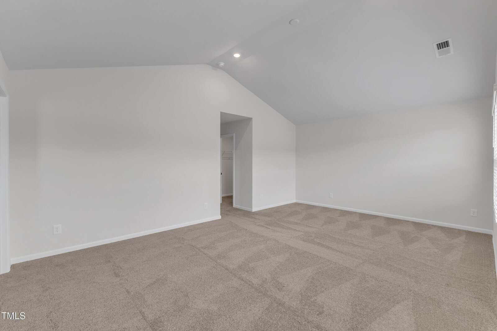 Empty room with light beige carpet, white walls, a vaulted ceiling, and a doorway leading to another area; overhead lighting and a ceiling vent are visible.