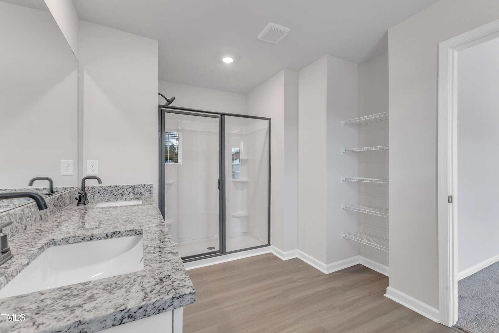A modern bathroom with a double sink vanity, granite countertop, a glass-enclosed shower, built-in wall shelves, and wood-look flooring. An open doorway leads to a carpeted room.