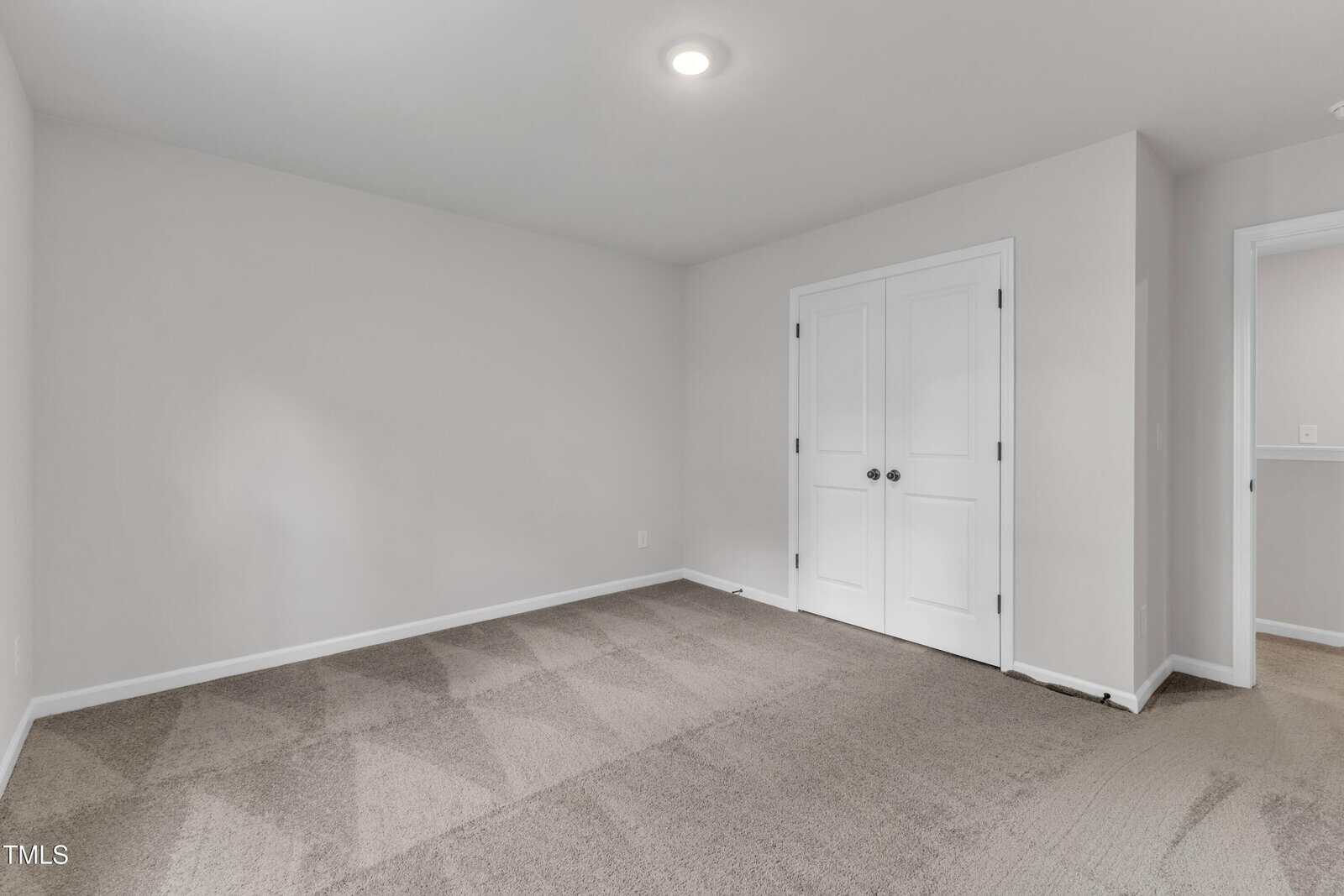 Empty room with beige carpet, white walls, a ceiling light, double white closet doors, and an open doorway leading to a hallway.
