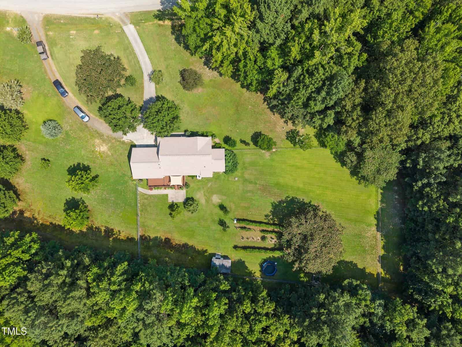 Aerial view of a house with a light-colored roof surrounded by green grass, trees, a driveway with parked cars, a backyard garden area, and a wooded area bordering the property.