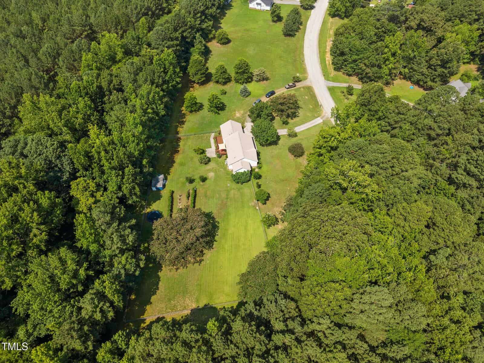 Aerial view of a house surrounded by large grassy lawns and dense trees, with a curved road and a few cars nearby; other houses and open spaces are visible in the surrounding area.
