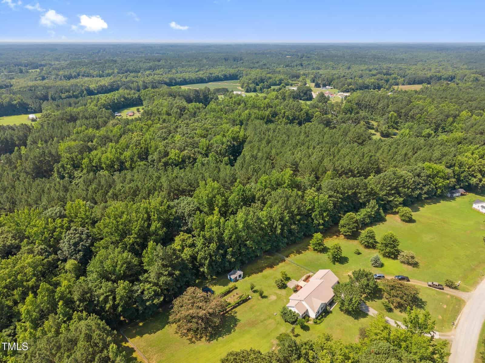 Aerial view of a house with a large yard surrounded by trees and forested areas, with a few neighboring homes visible and expansive greenery extending into the distance under a blue sky.