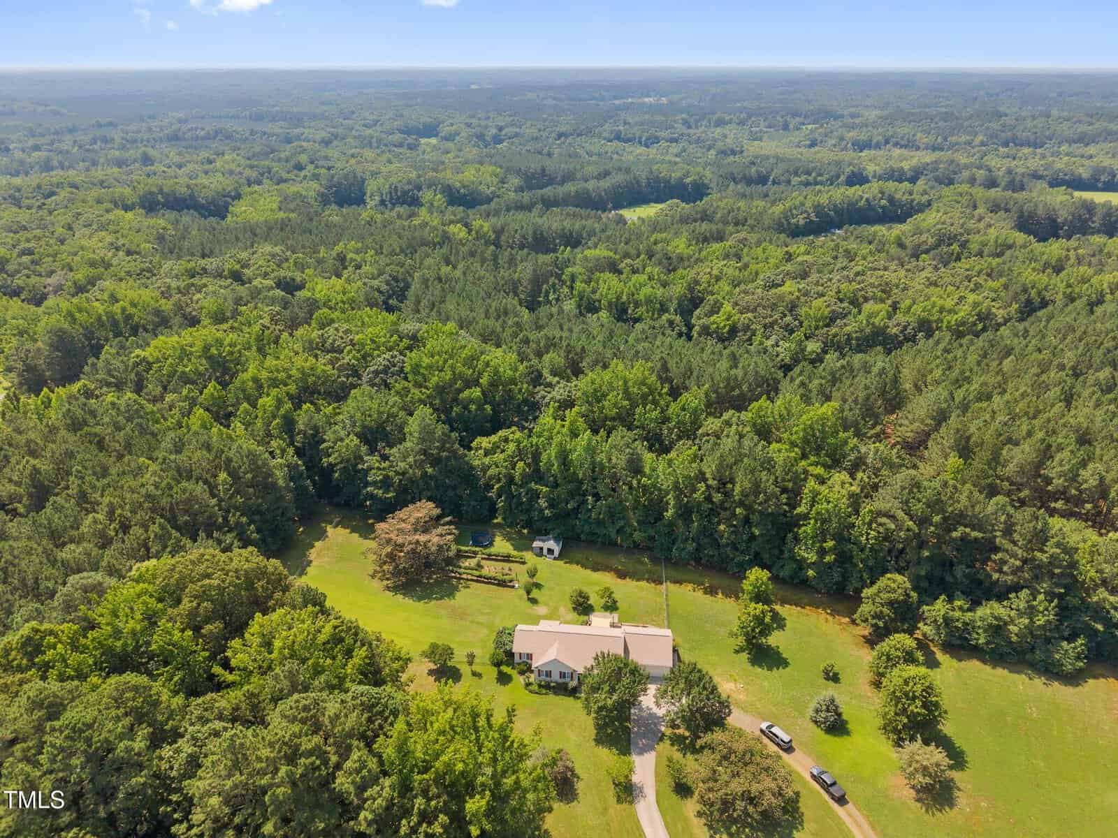 A large house with a white roof sits on a spacious, grassy lot surrounded by dense forest. A long driveway leads to the house, and several outbuildings and trees dot the property. Rolling hills and trees fill the background.