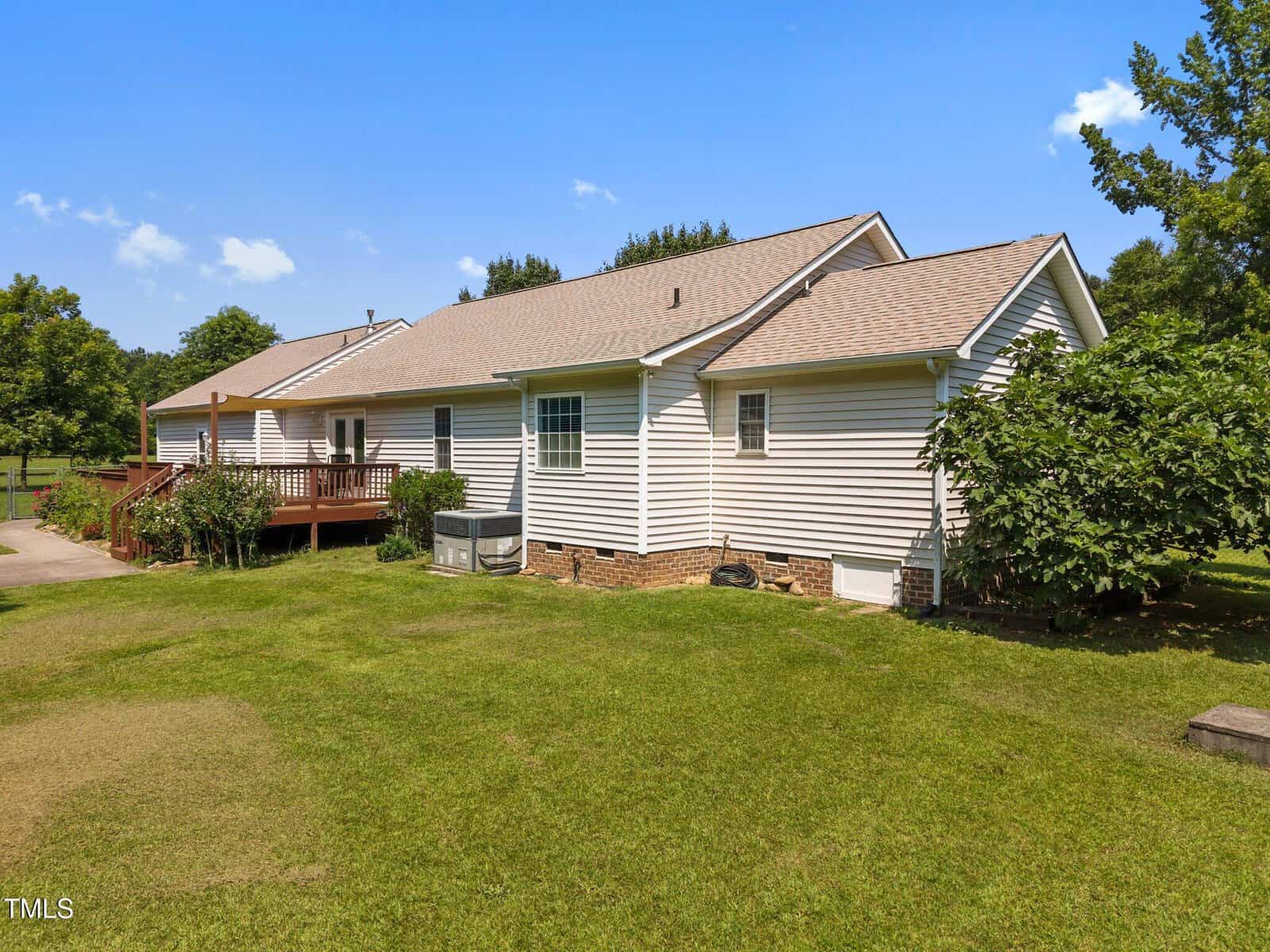 Single-story house with beige siding, a shingled roof, a wooden deck, an HVAC unit outside, and a green lawn with some trees and shrubs, under a clear blue sky.