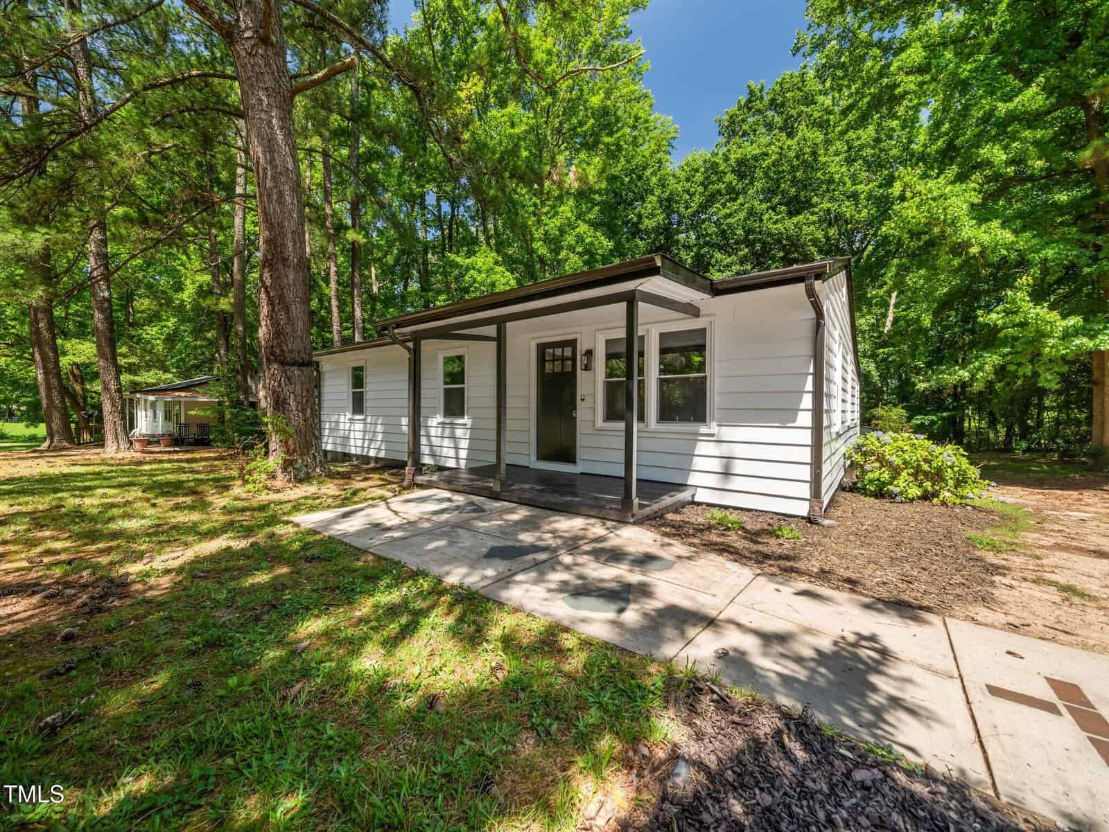 A small white house with a dark front door, flat roof, and covered porch sits surrounded by tall trees and lush green grass on a sunny day. A sidewalk leads up to the entrance.