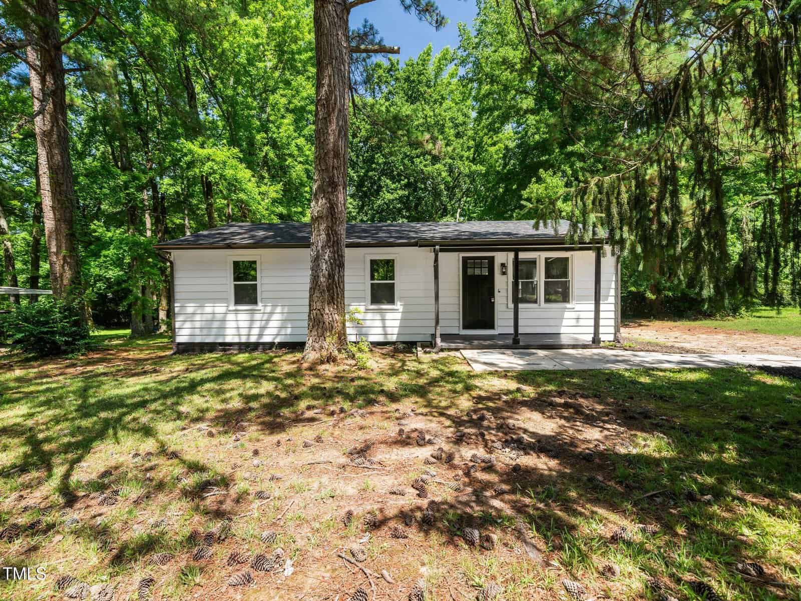 Small white single-story house with black trim and a dark front door, surrounded by tall green trees and a grassy yard, shown on a sunny day. A concrete path leads to the entrance.