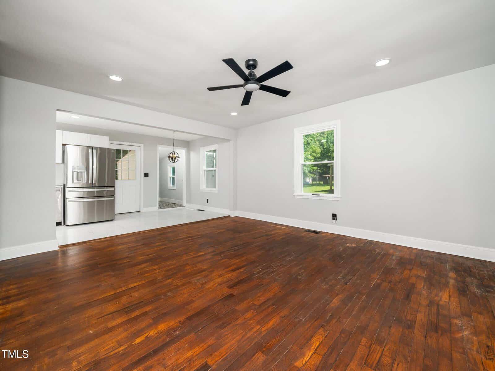 Spacious living room with dark hardwood floors, white walls, a black ceiling fan, and recessed lighting, open to a modern kitchen with stainless steel appliances and large windows letting in natural light.