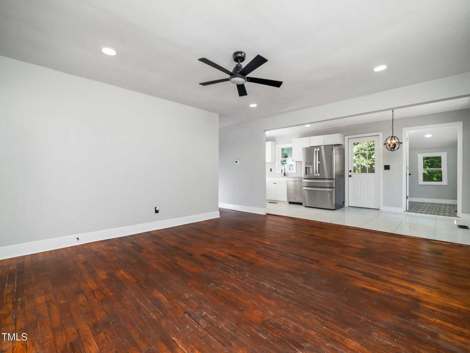 Spacious, modern living room with dark wood floors, white walls, ceiling fan, and recessed lighting. Open layout leads to a bright kitchen with stainless steel appliances and tiled flooring.
