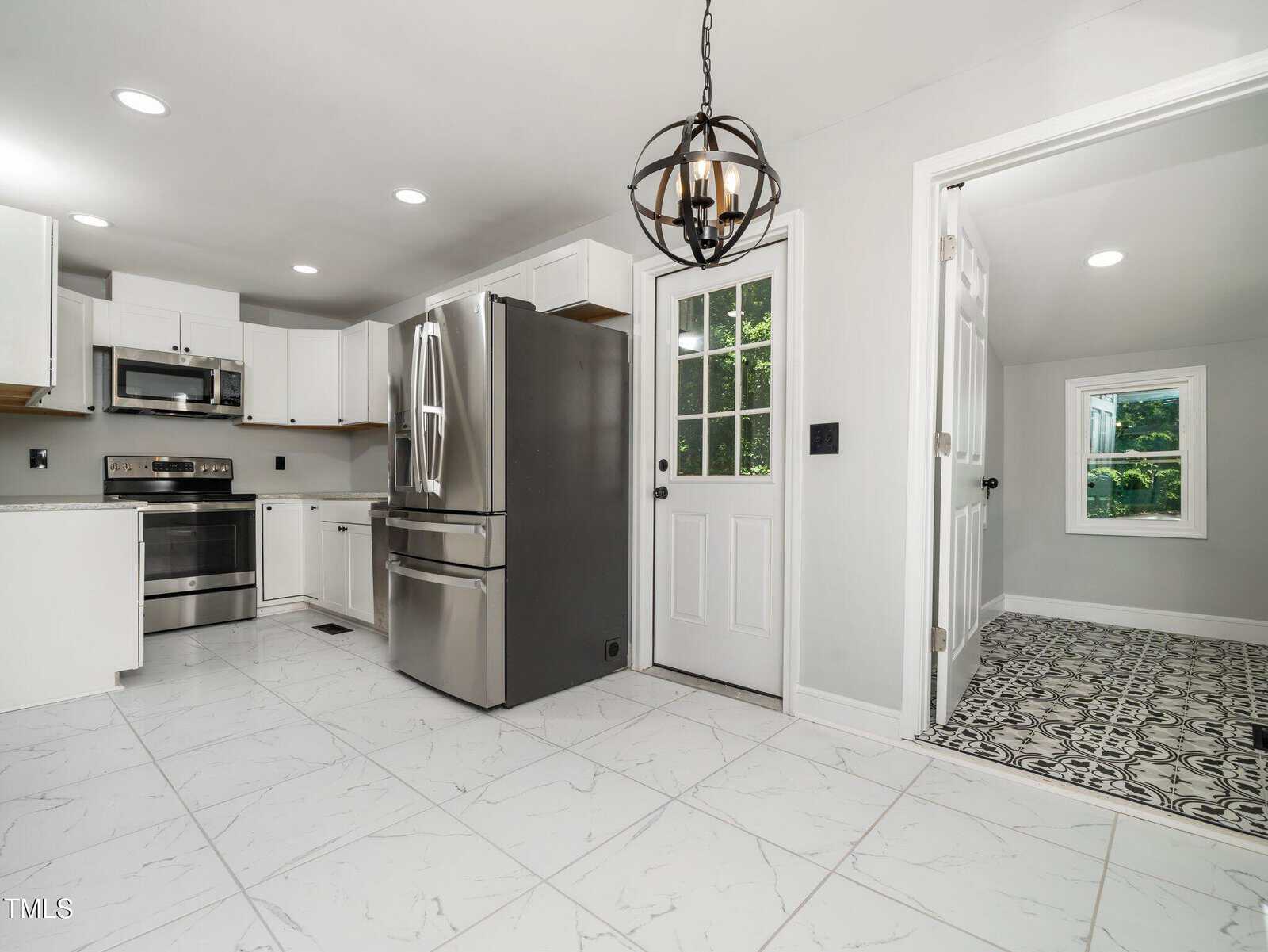 Modern white kitchen with stainless steel appliances, white cabinets, marble-patterned tile floor, a pendant light fixture, and adjoining laundry room with patterned floor tiles and a window.