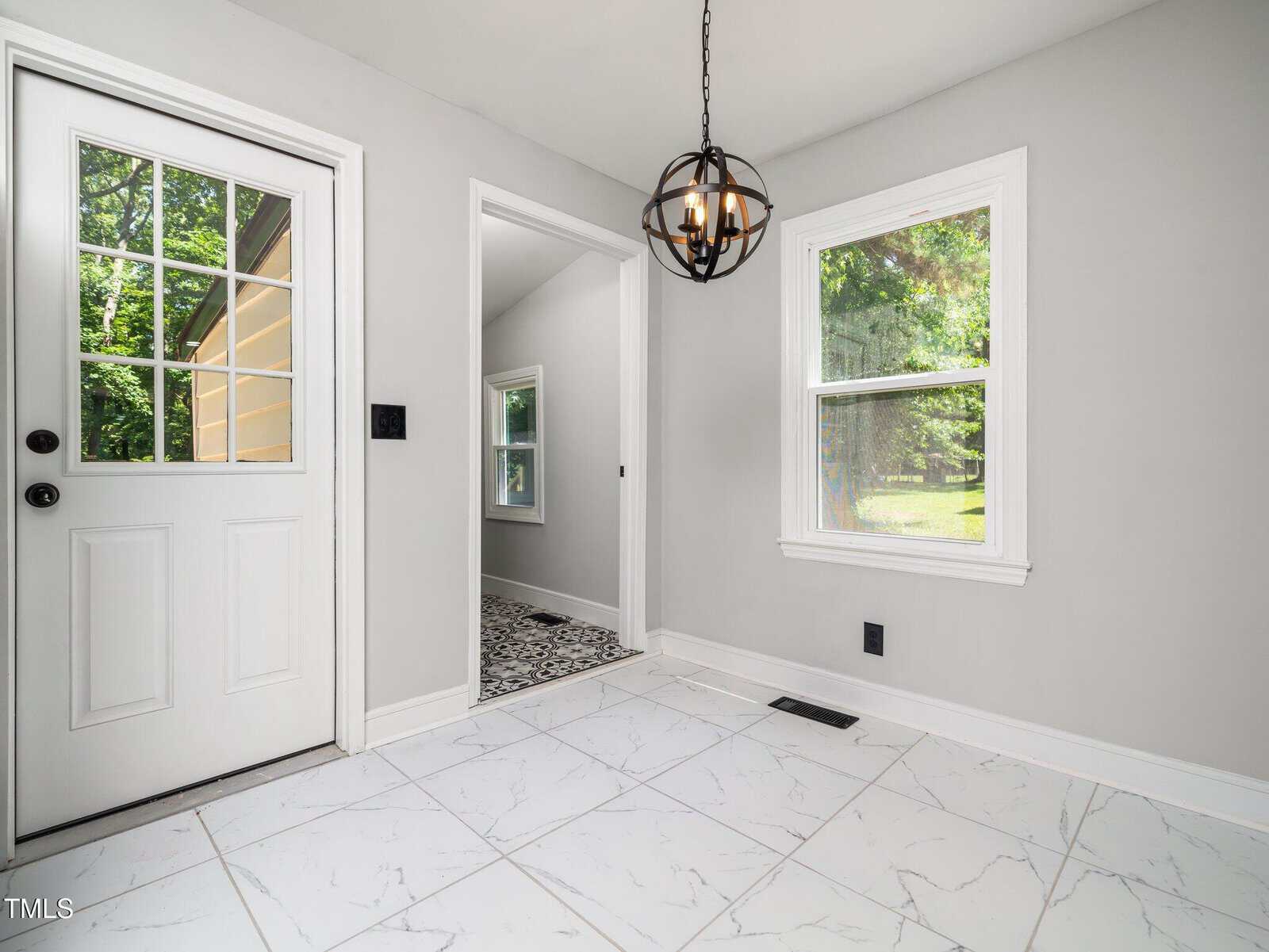 A bright room with white marble tile flooring, light gray walls, two windows, a black light fixture, and a door with glass panels leading outside. An open doorway leads to another room with patterned flooring.
